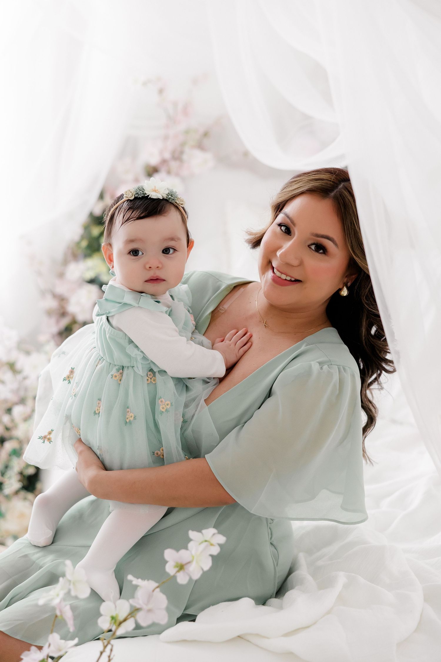 Parent and baby in matching mint green outfits pose together with white flowers in a bright studio setting.