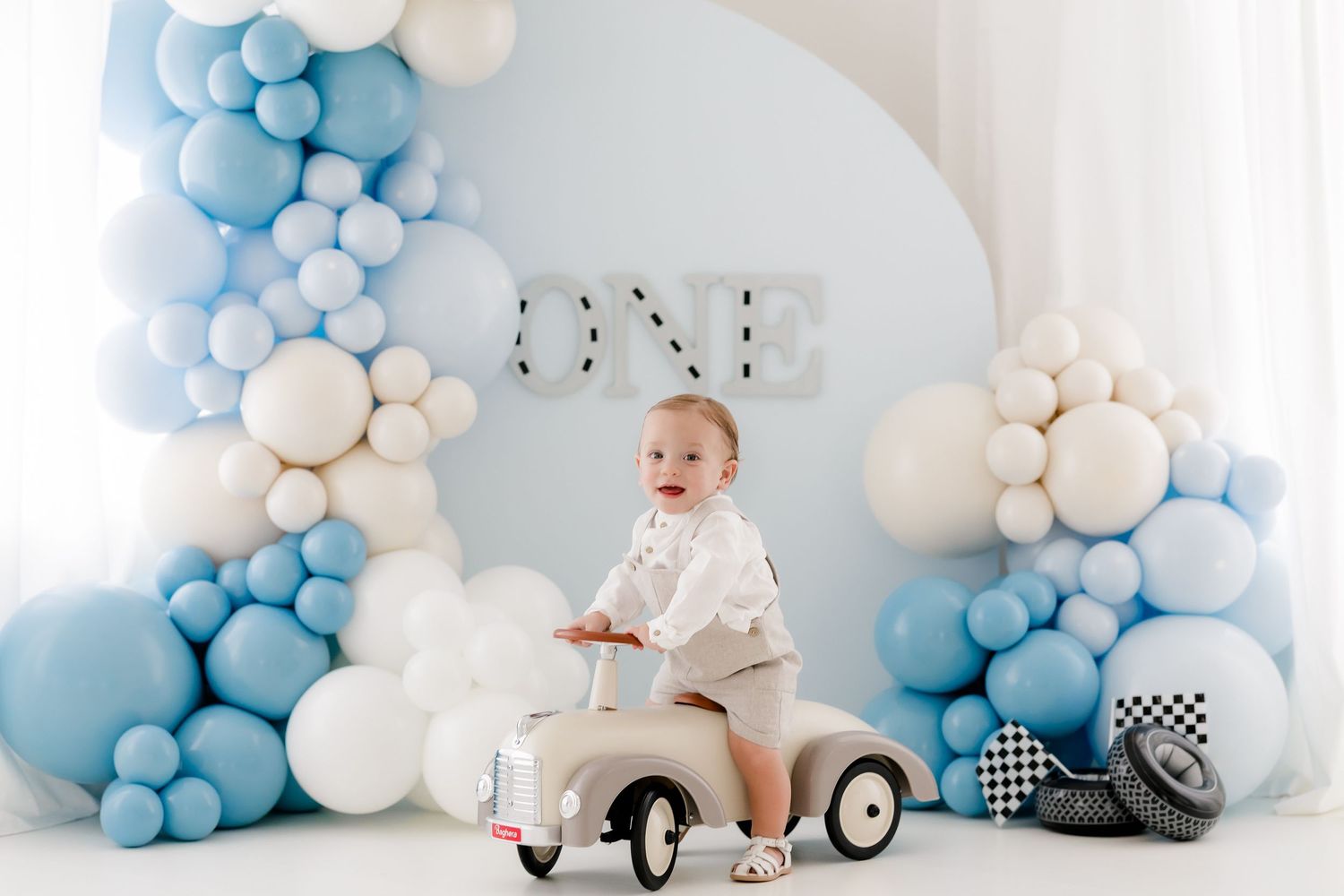 First birthday celebration setup with vintage toy car, blue and white balloon arch, and soft pastel decorations.