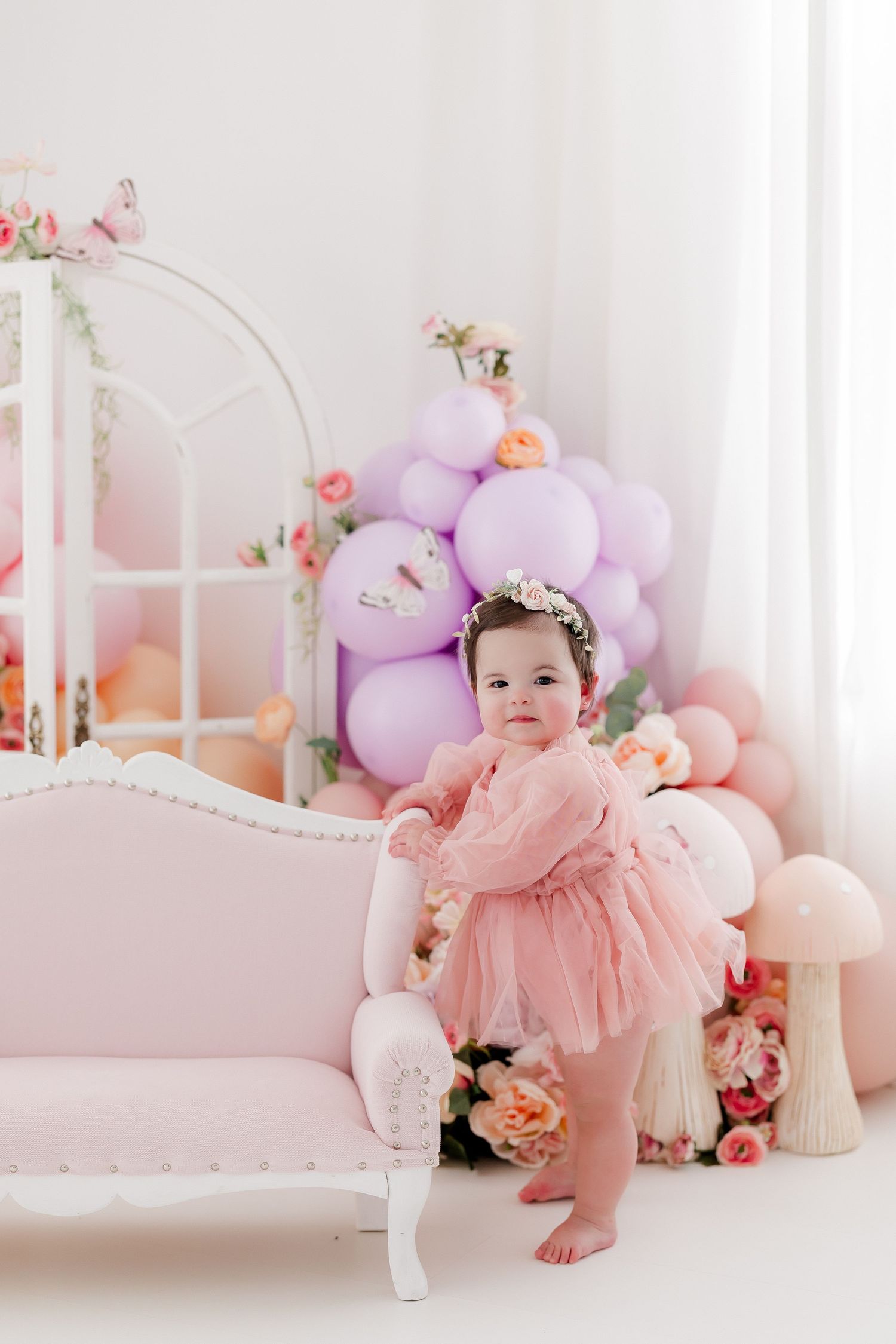 Little one in pink tulle dress poses on white bench with floral decor and balloons.