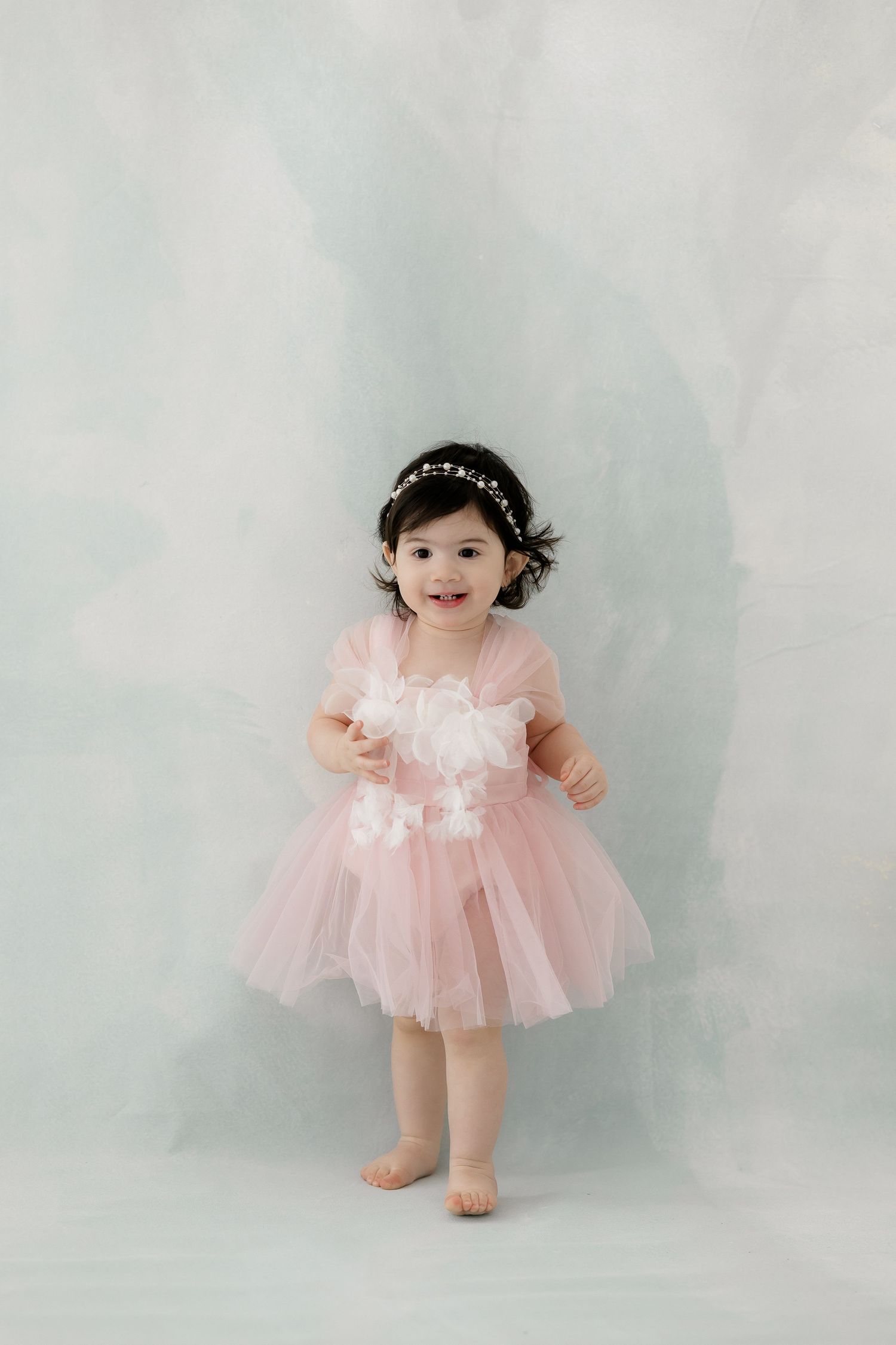 A toddler wearing a pink tulle dress and sparkly headband stands smiling against a light gray background.