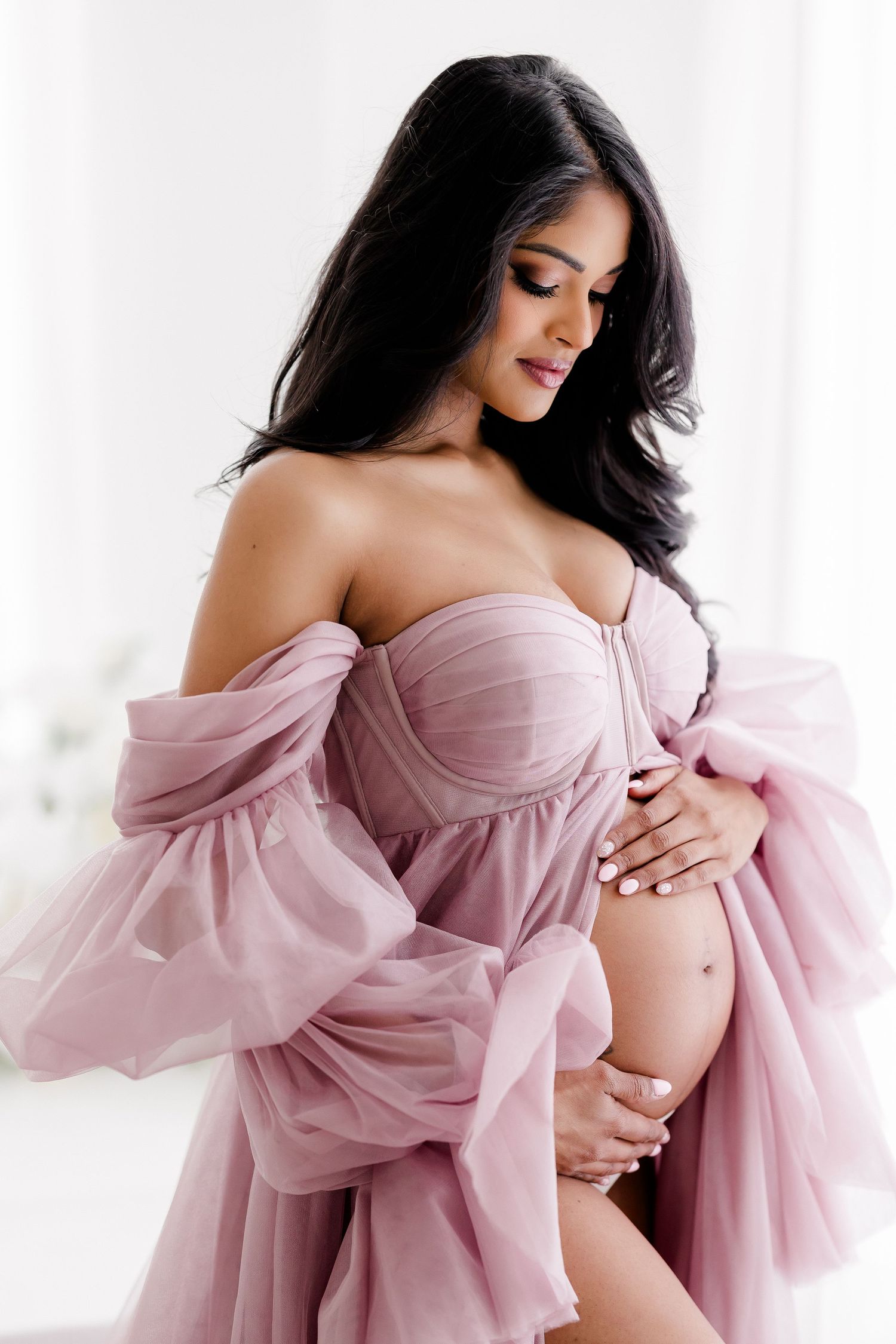 A maternity photo series showing a woman in a flowing pink off-shoulder gown against a bright white backdrop.