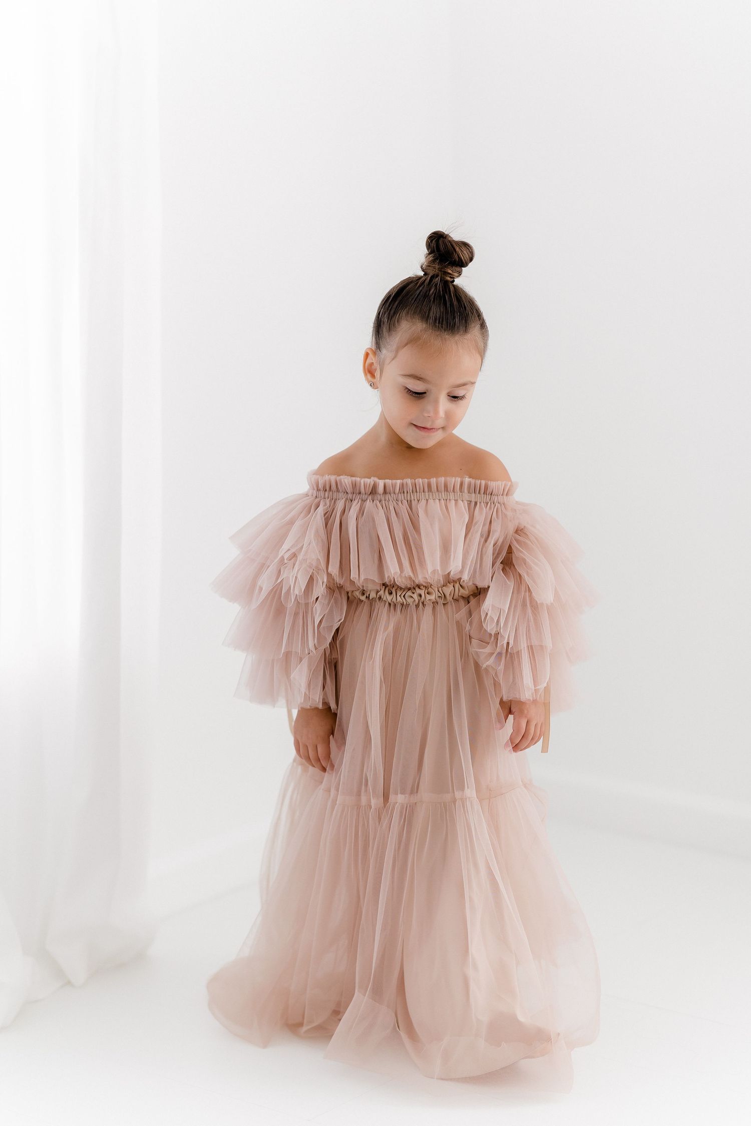 Child wearing off-shoulder blush tulle gown poses elegantly in studio setting.
