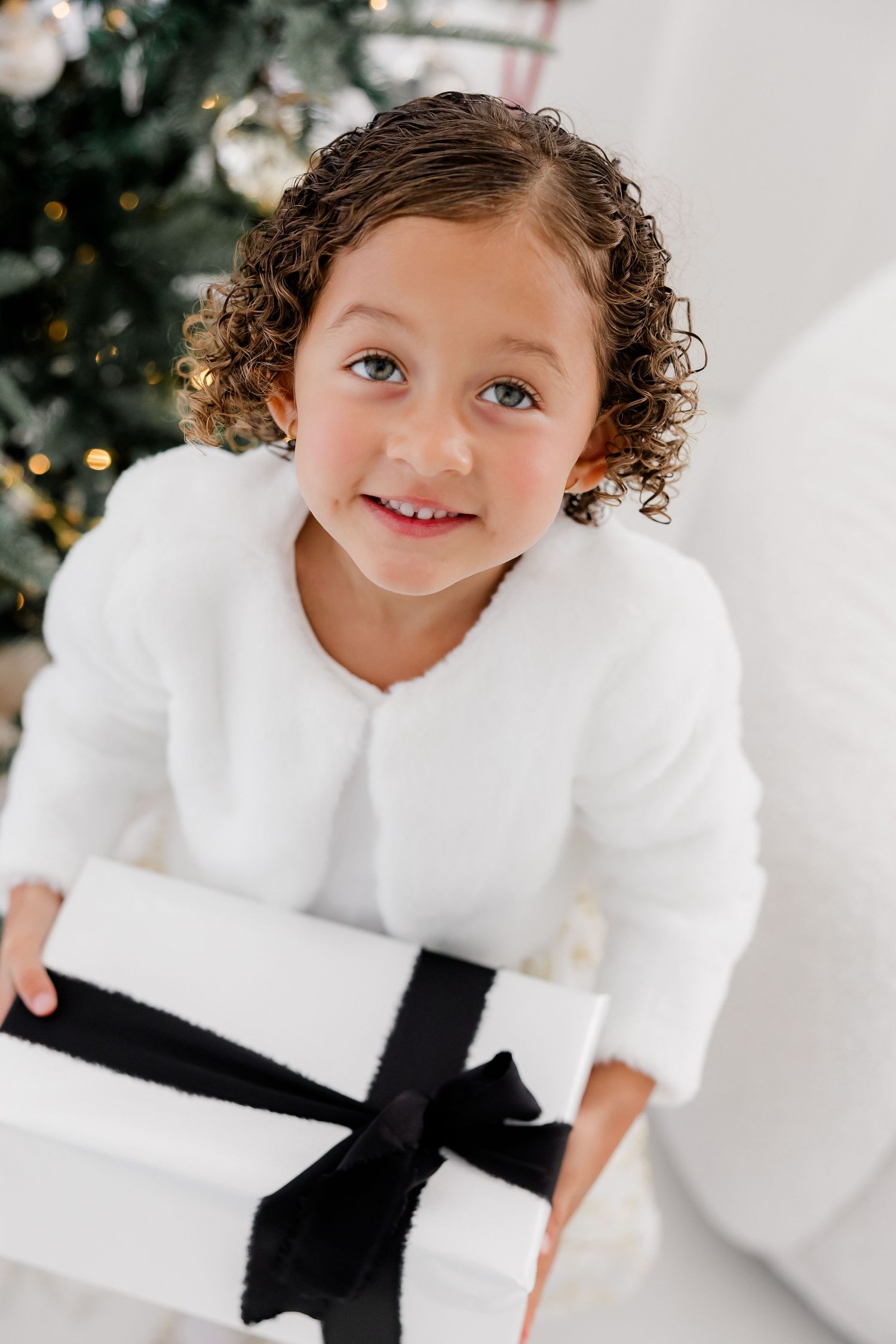 Child holding a white gift box with black bow in front of Christmas tree decorations.