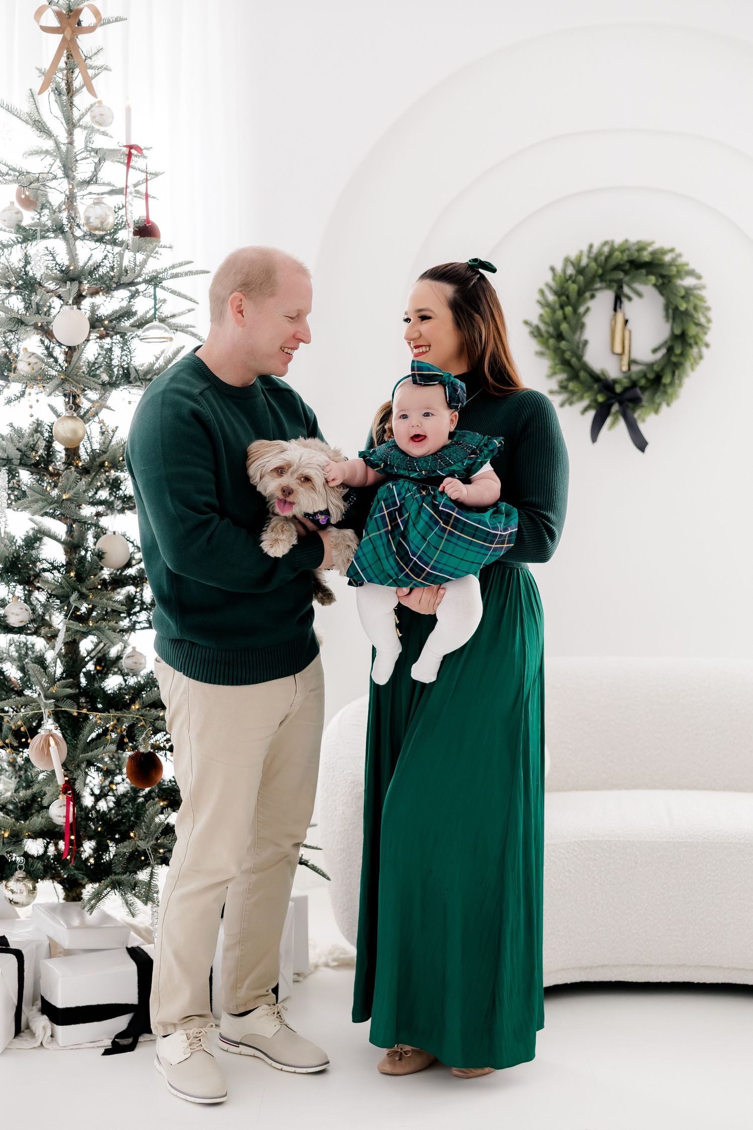 Family in coordinating green outfits poses with dog by Christmas tree.