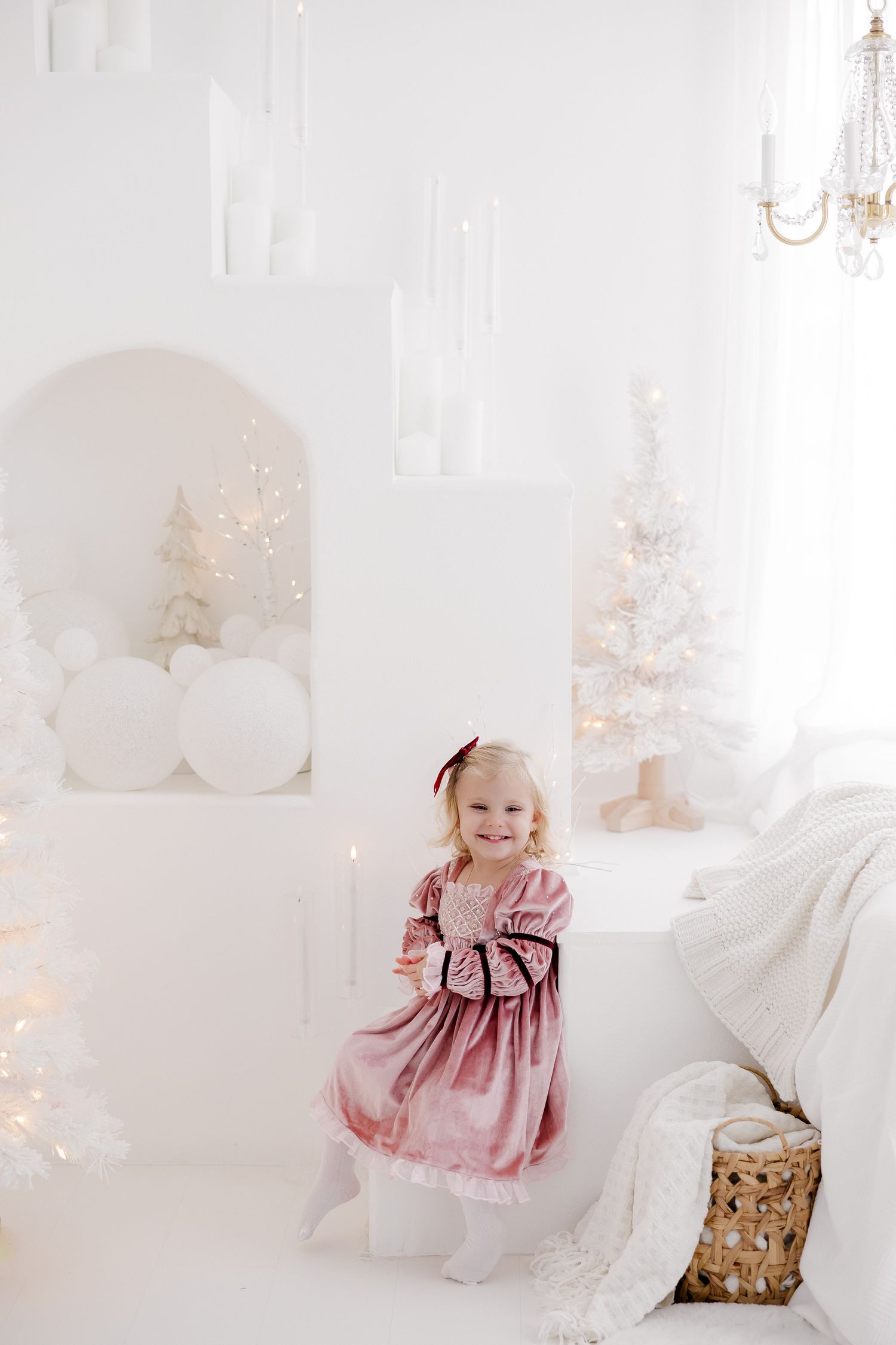 Toddler in pink dress sitting in elegant white studio with holiday lights and decor.