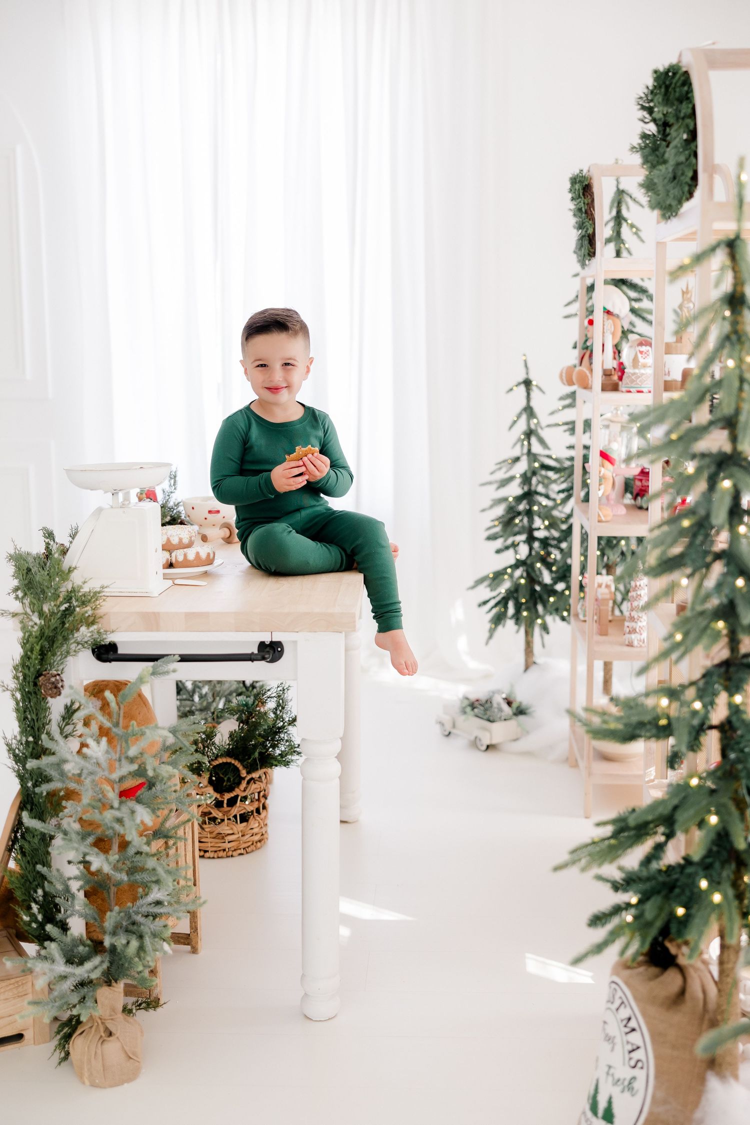 Child in green outfit sitting on white counter with Christmas decorations and miniature trees.