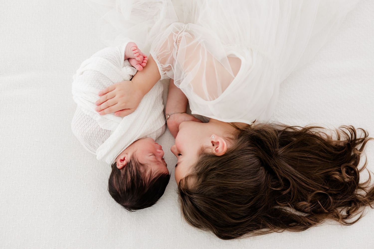 Dark-haired person lying on white fabric surface wearing white clothing against light background.
