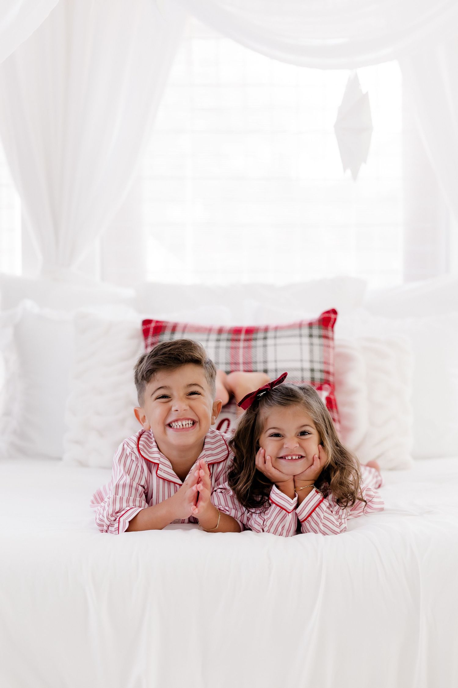 Two smiling children in matching red striped pajamas lying on a white bed with plaid pillows.