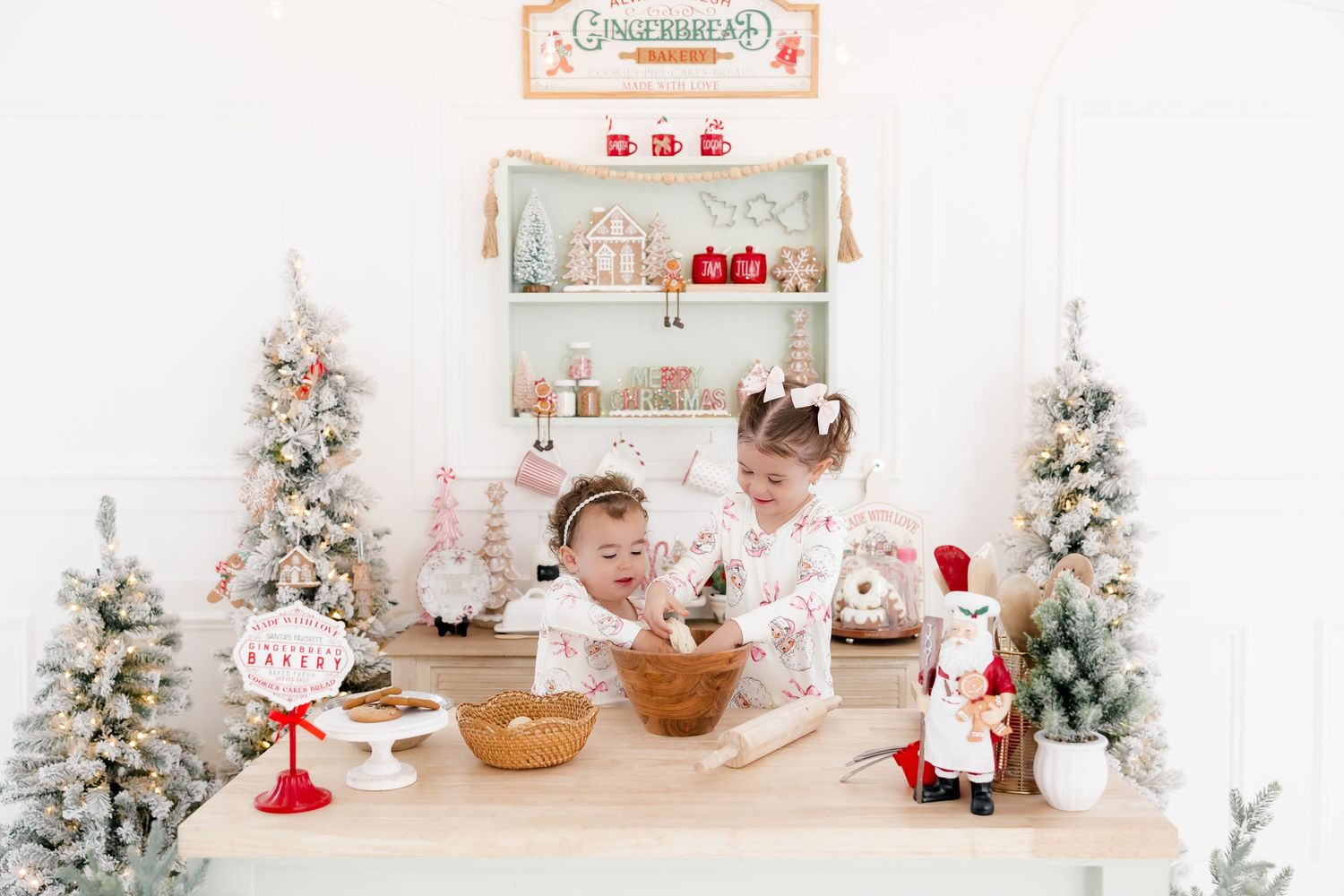 Two young girls bake together at a festive Christmas table with snowy trees, nutcracker, and gingerbread shop backdrop.