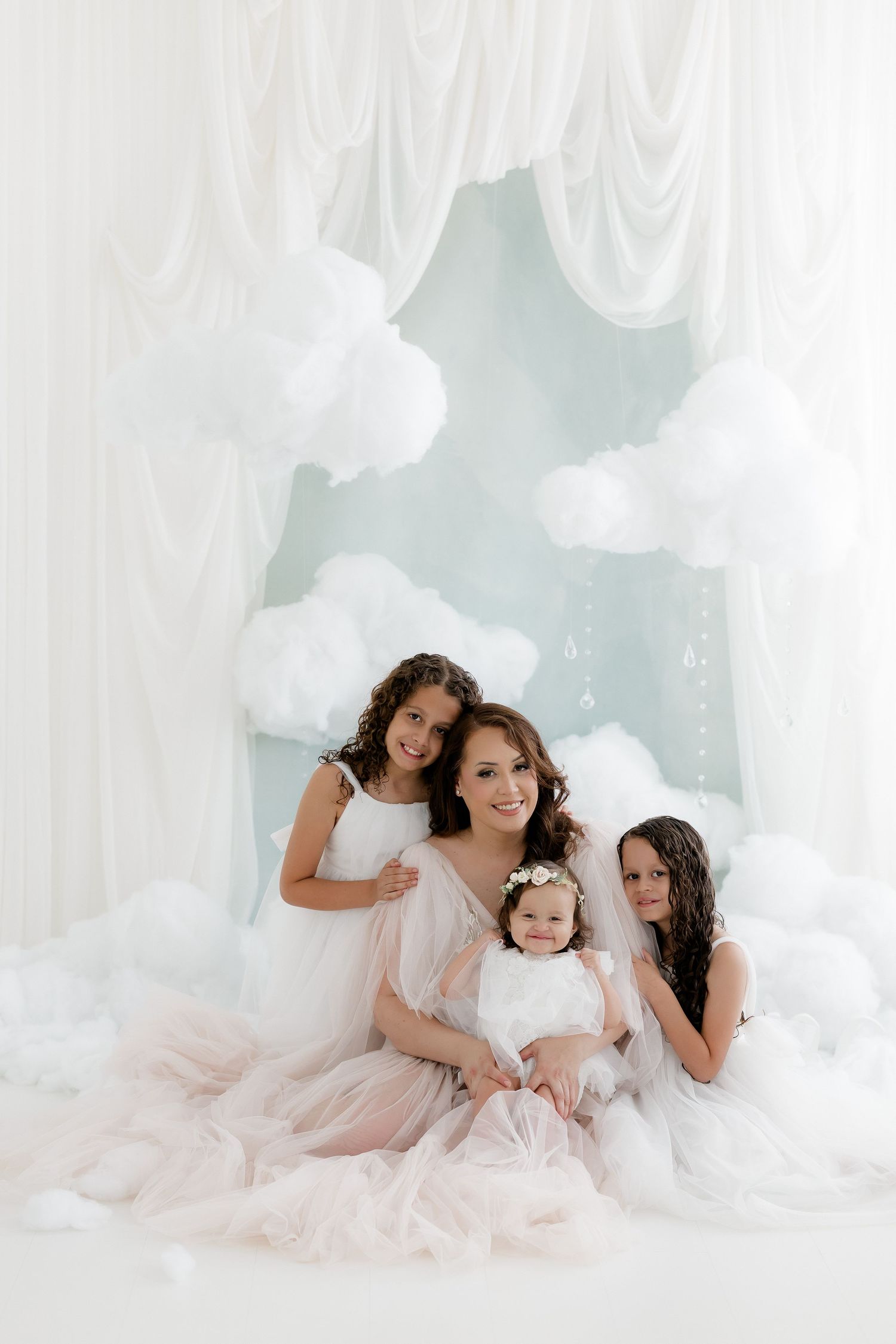 Mother poses with three young daughters in white dresses in elegant cloud-themed photo studio.