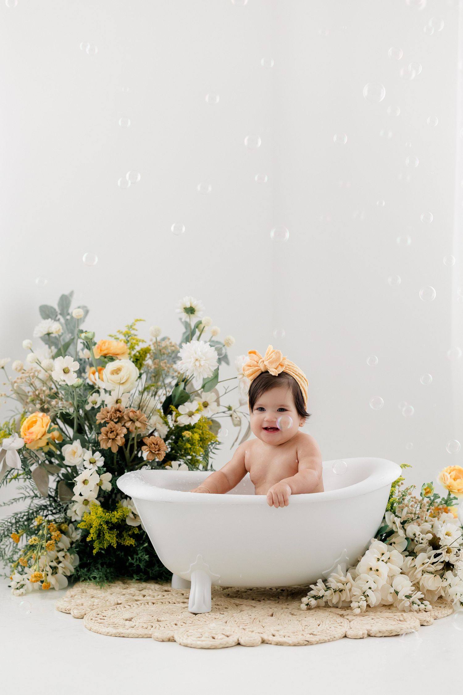 Baby sitting in white bathtub surrounded by yellow and white flowers and greenery on beige backdrop at a New Jersey photography studio