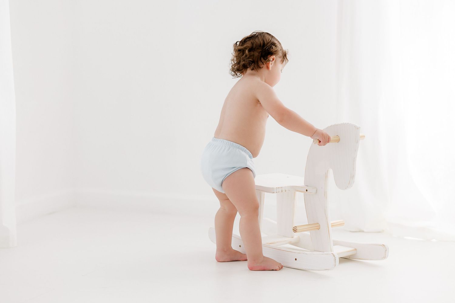 A toddler in a diaper stands next to a white wooden stool in a bright, minimalist setting.