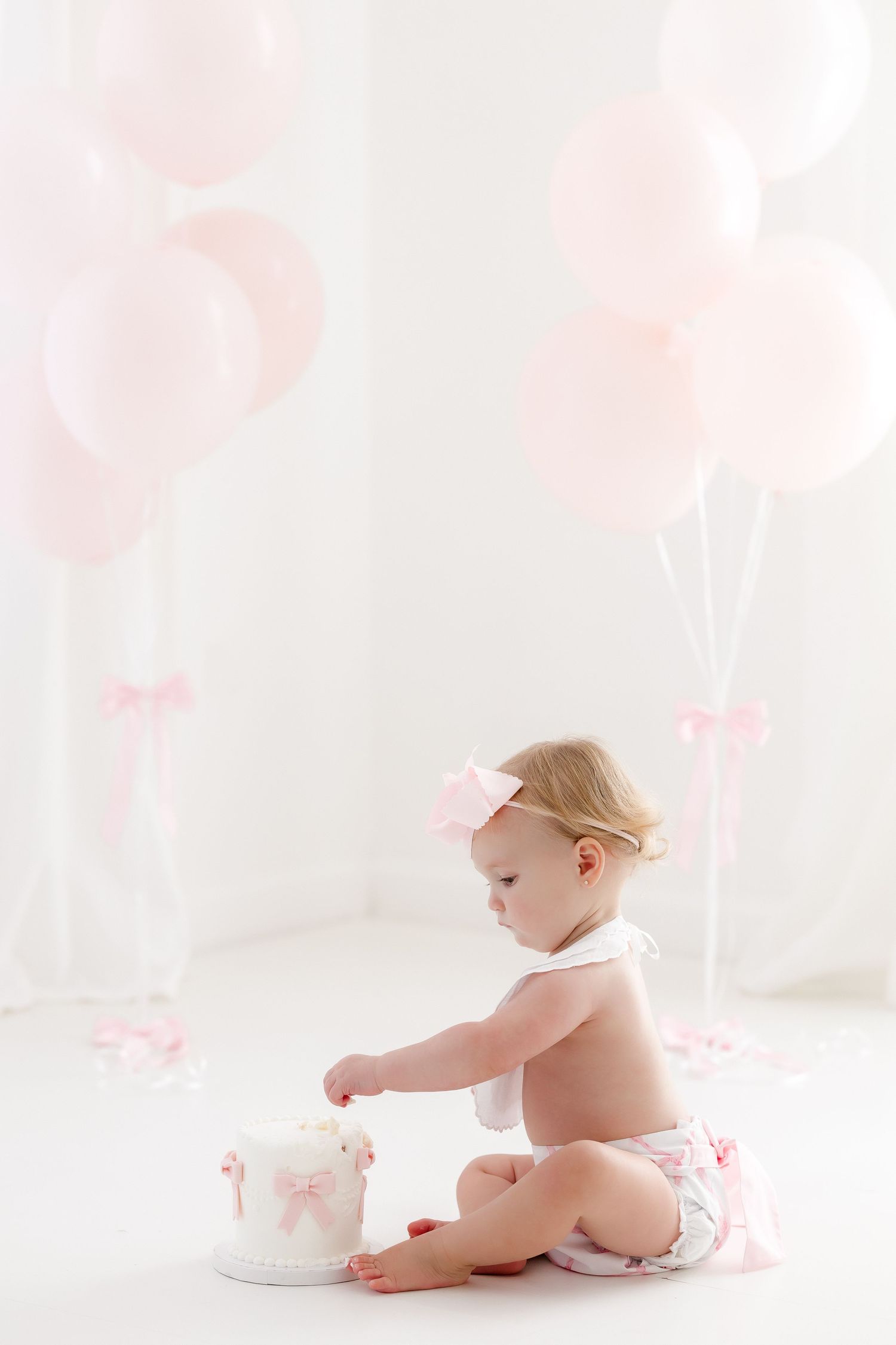Baby in white diaper and pink tutu sits on floor with soft pink floral background and white decor elements.