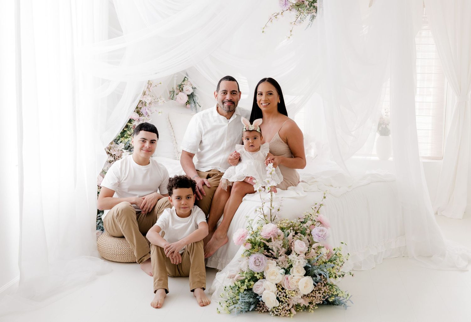 A family in white outfits sits together on a couch for a series of bright, airy indoor portrait photos.