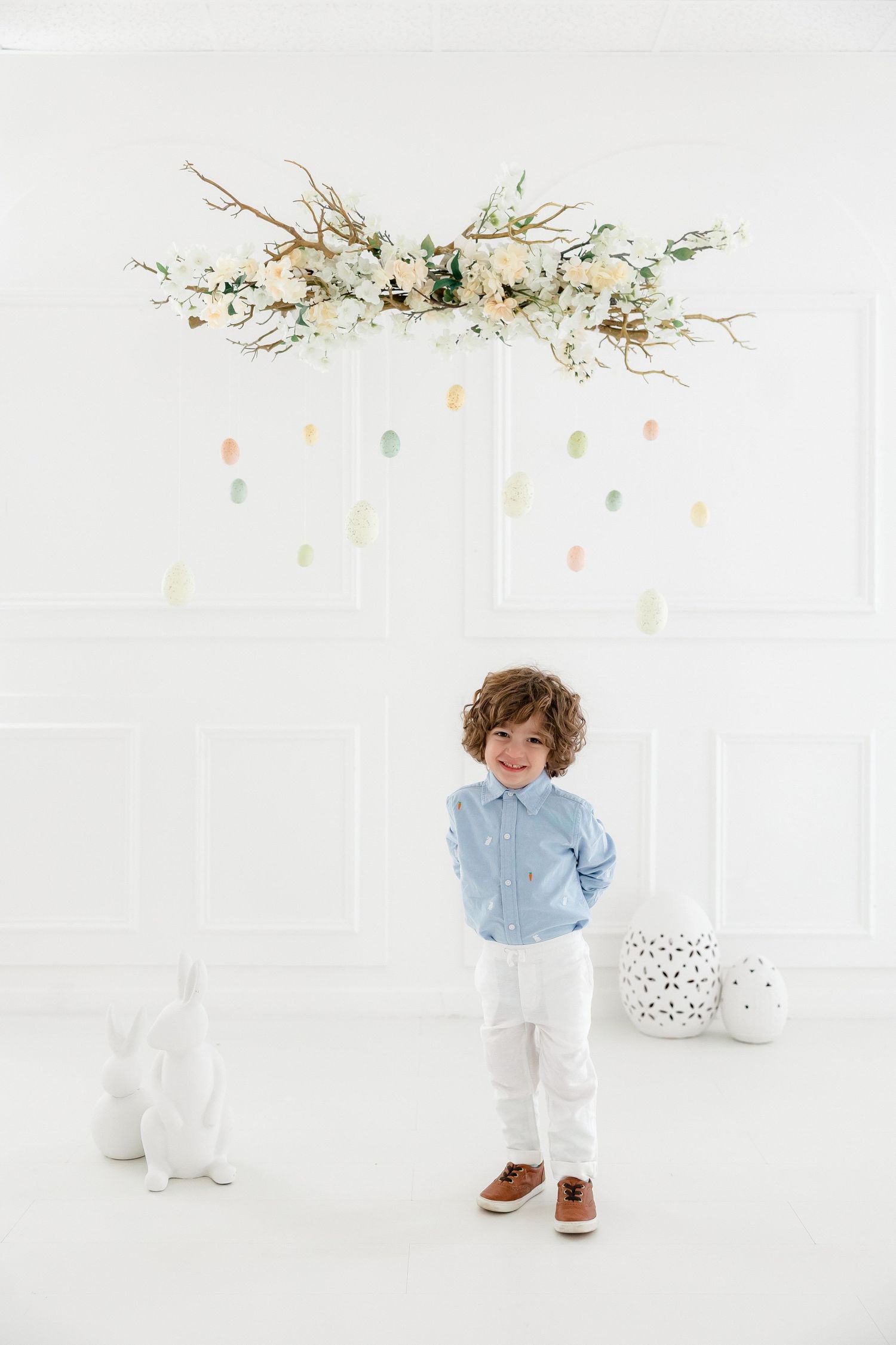 Small child wearing light blue shirt and white pants stands against white backdrop with floral decorations above.