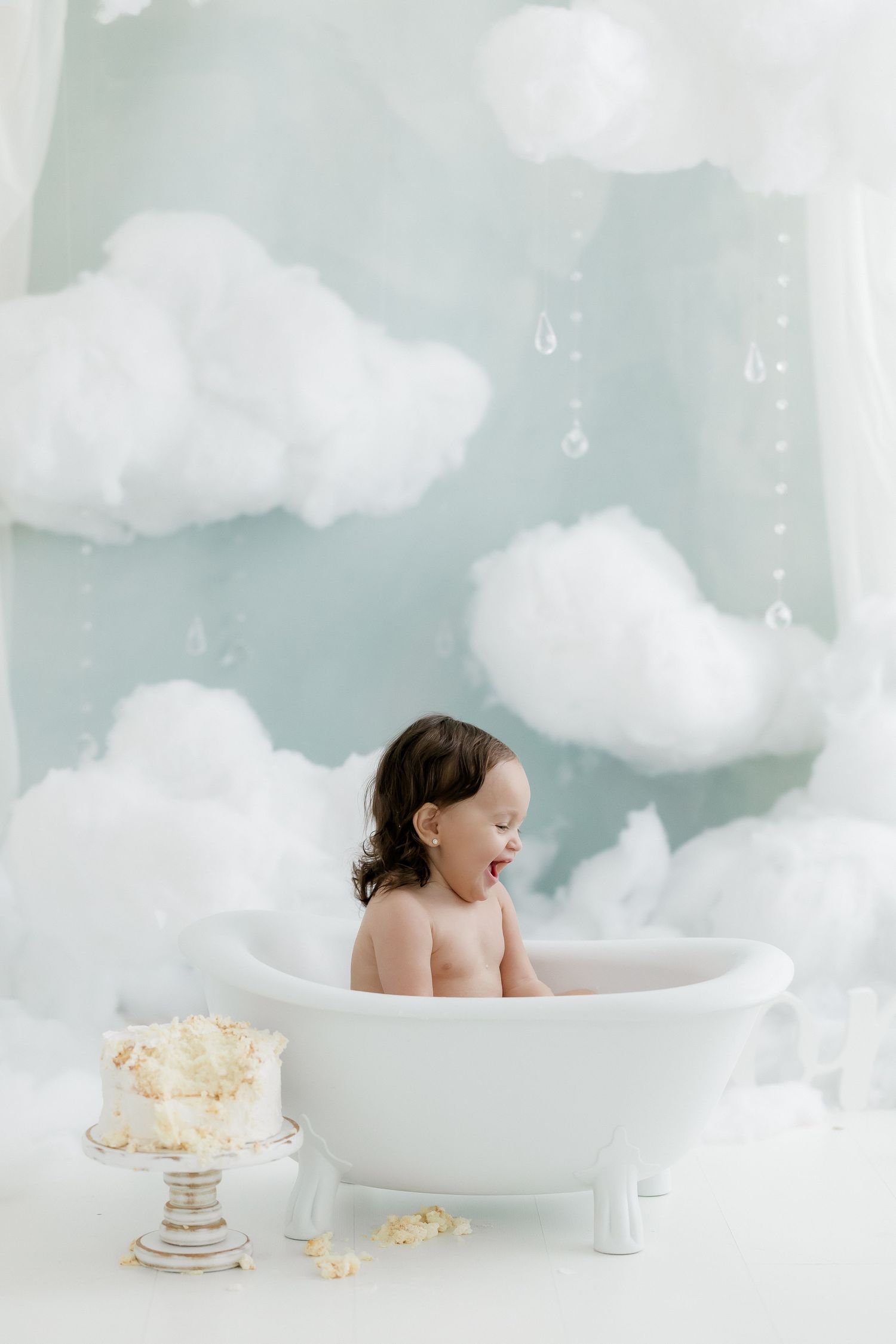 Smiling baby sitting in a white clawfoot bathtub with cloud decorations and a smash cake nearby.