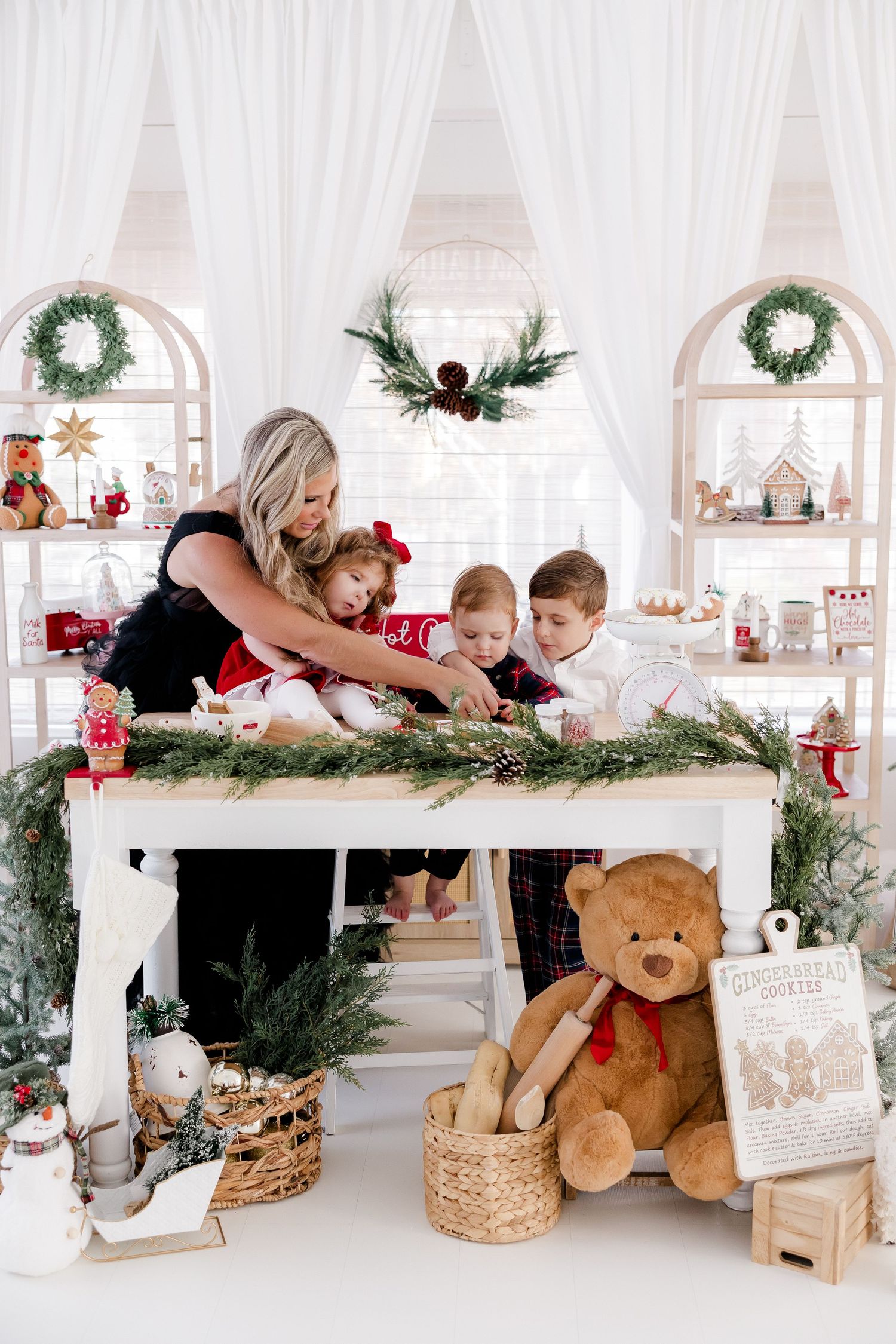 Holiday photo session setup with white decor, greenery and teddy bears on display shelves.