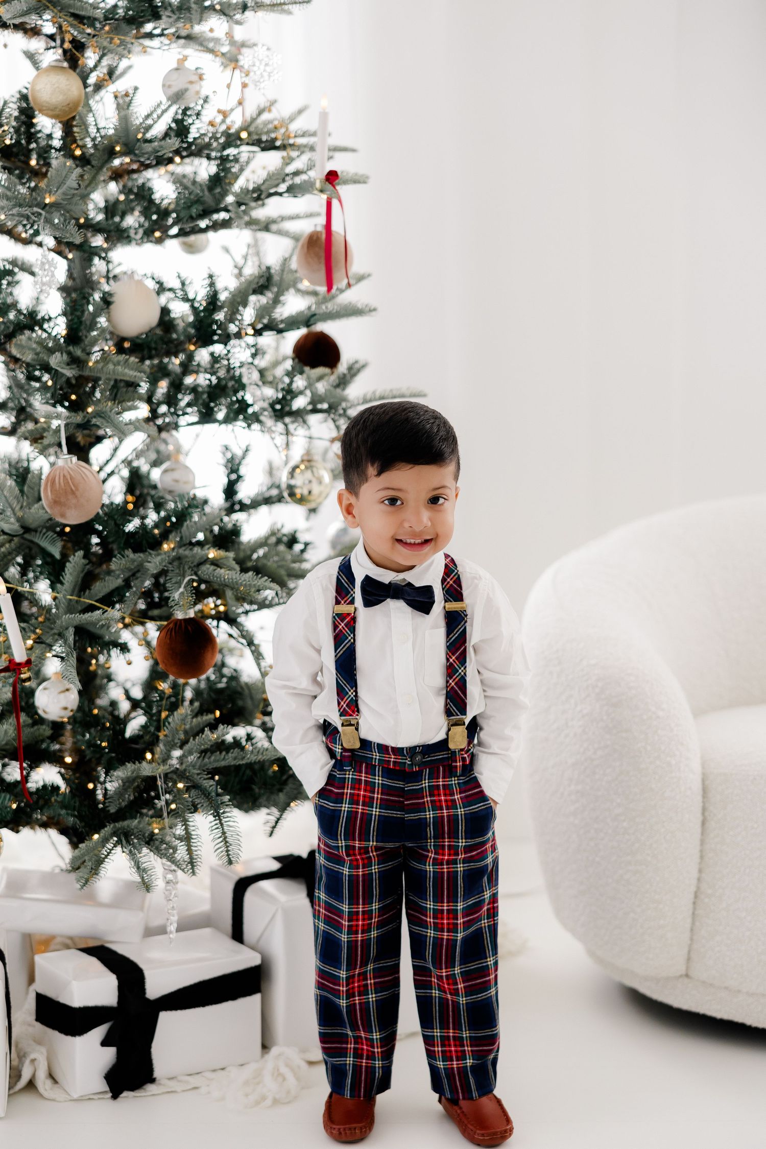 Toddler in festive plaid suspenders and bow tie poses by Christmas tree in white studio.
