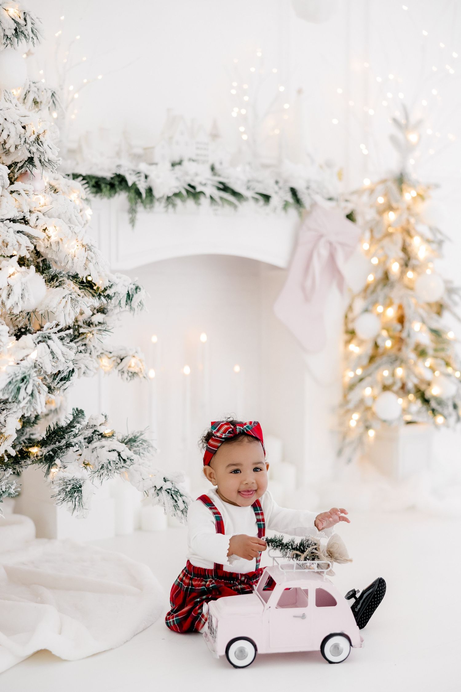 Child in red plaid outfit plays with pink toy car in winter wonderland studio setting.