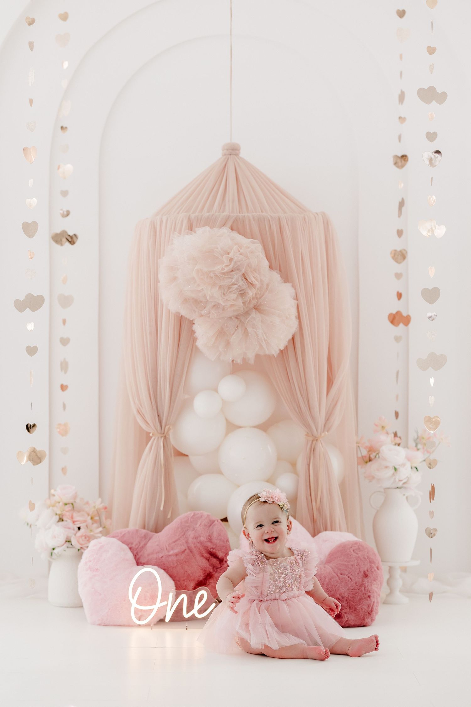 Pink birthday setup with tulle canopy, heart garlands, and neon 'One' sign.