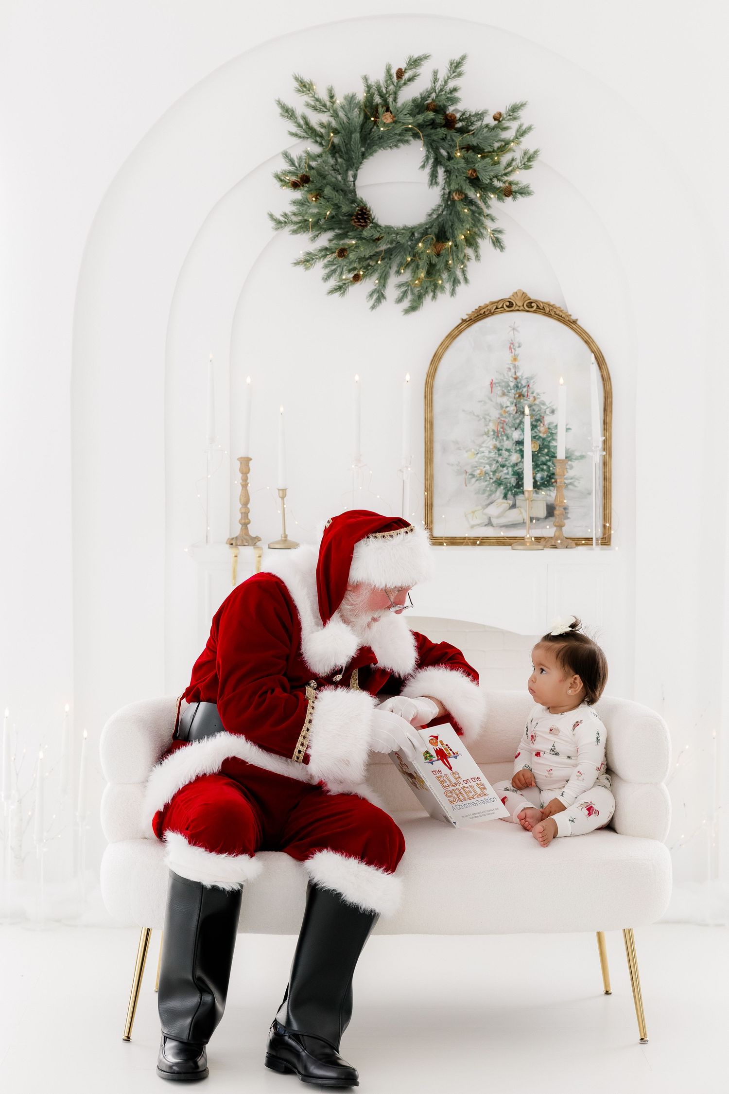 Santa Claus reading a book to a baby girl on a white sofa in a festive holiday setting with wreath decor.