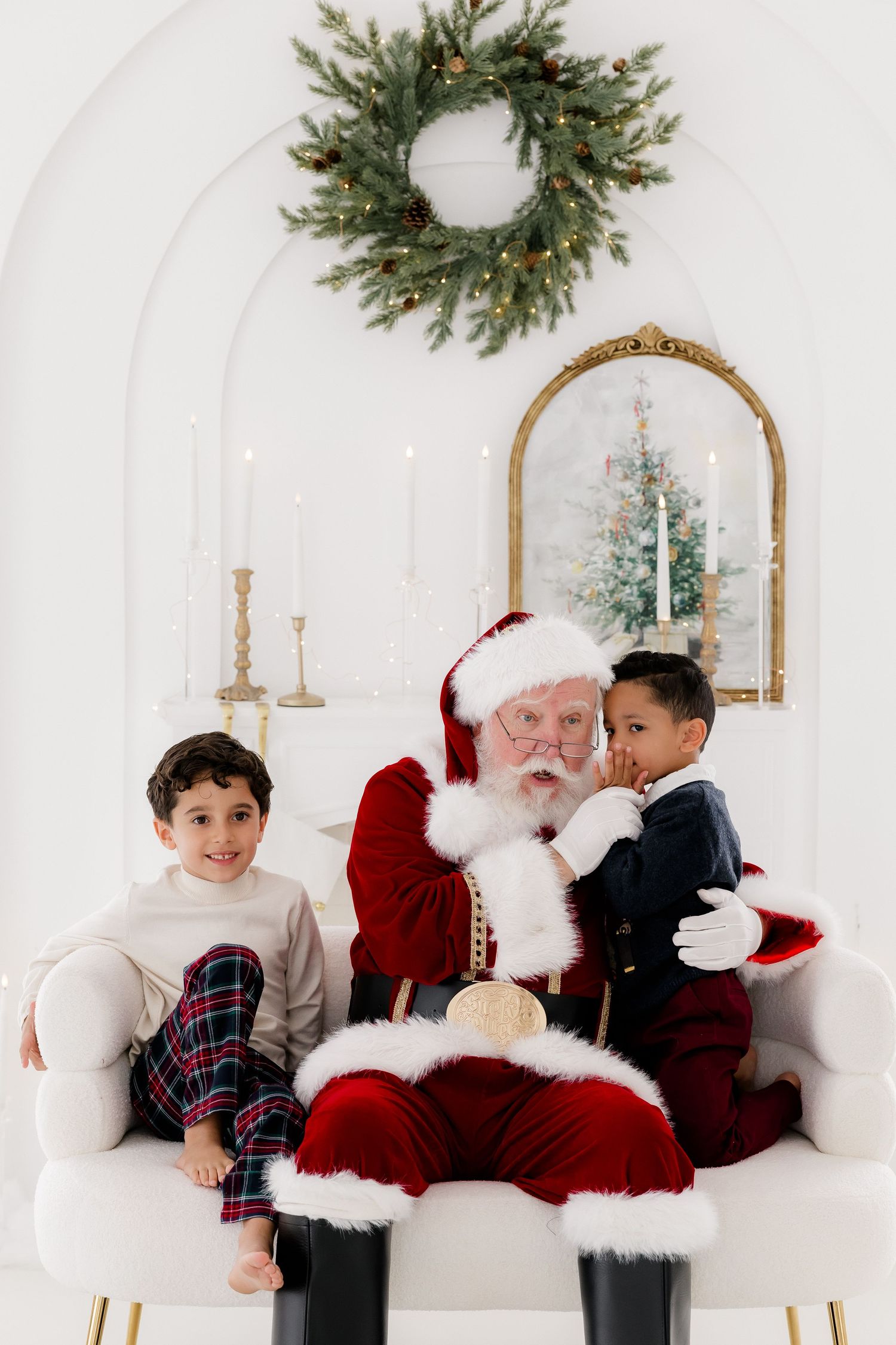 Two boys sit with Santa Claus on a white chair, surrounded by festive Christmas decor including a wreath and candles.