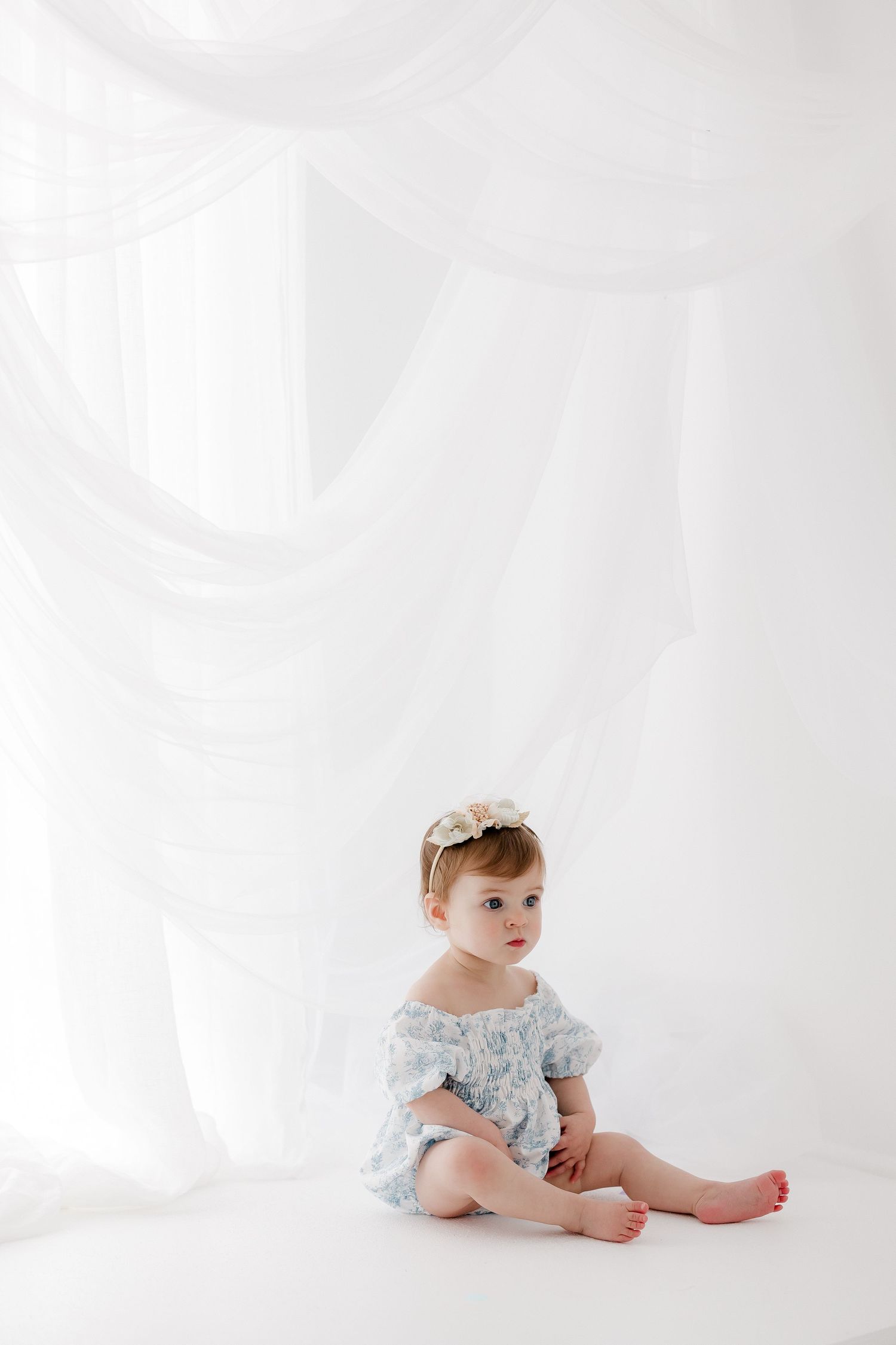 Baby wearing light blue floral dress sits on white backdrop in minimalist studio setting.