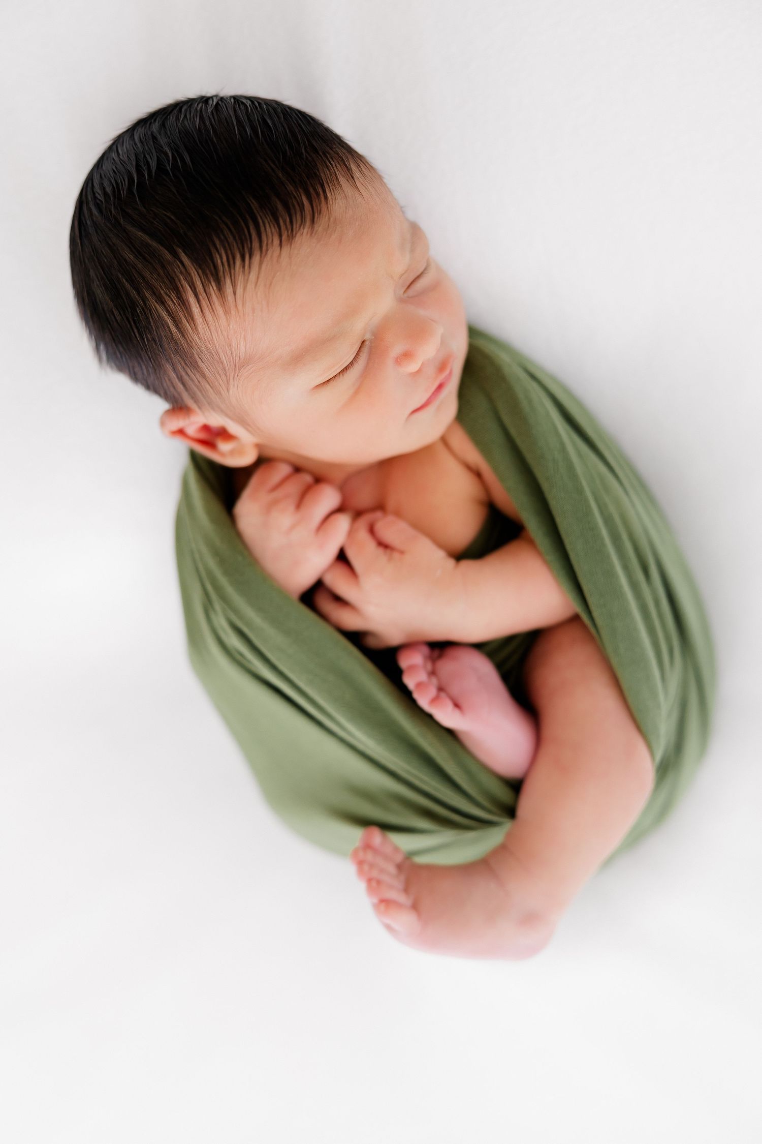 Sleeping newborn baby wrapped in olive green fabric against white background in minimalist style portrait series.