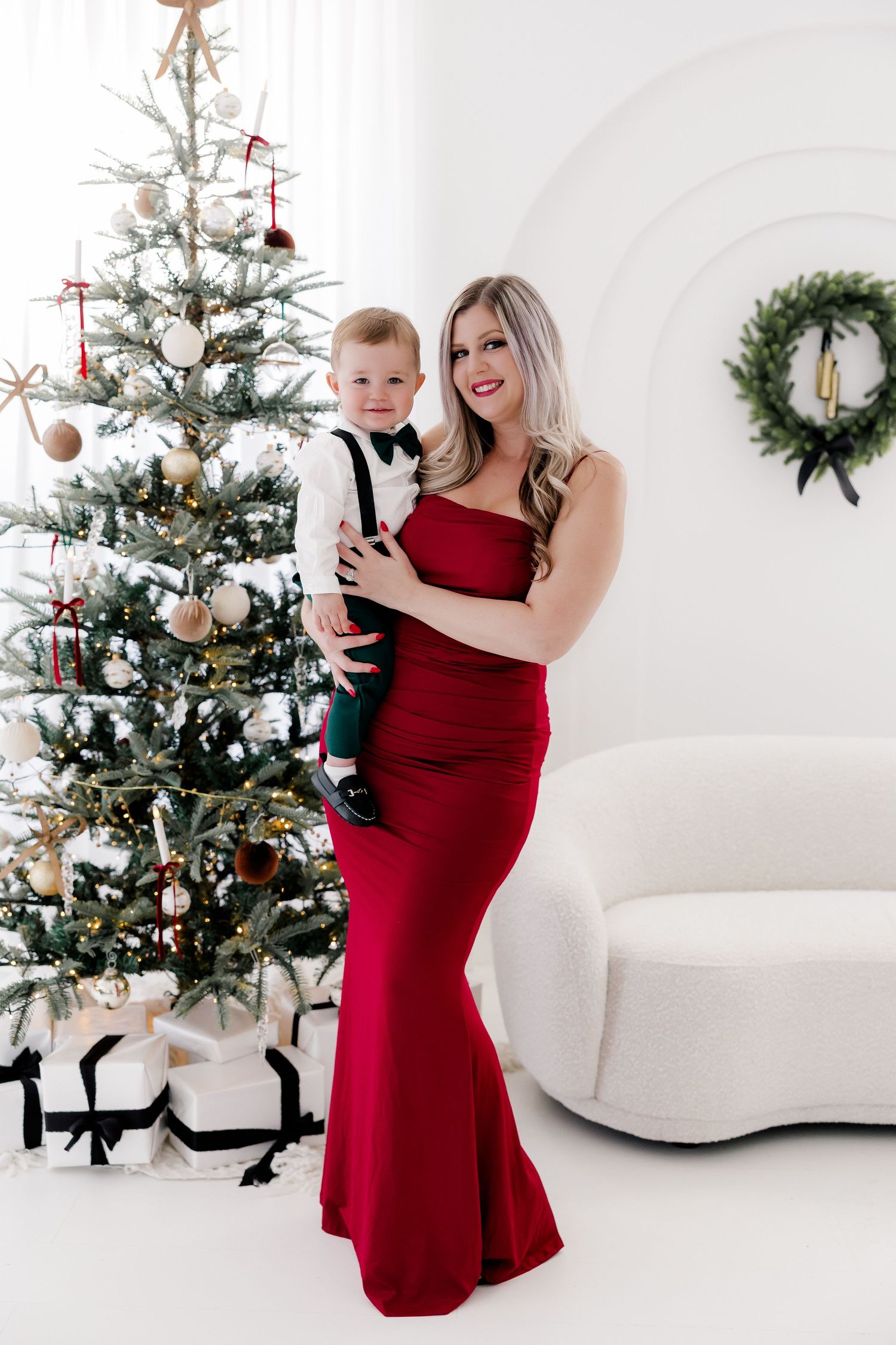Woman in red dress poses elegantly by decorated Christmas tree in white studio setting.