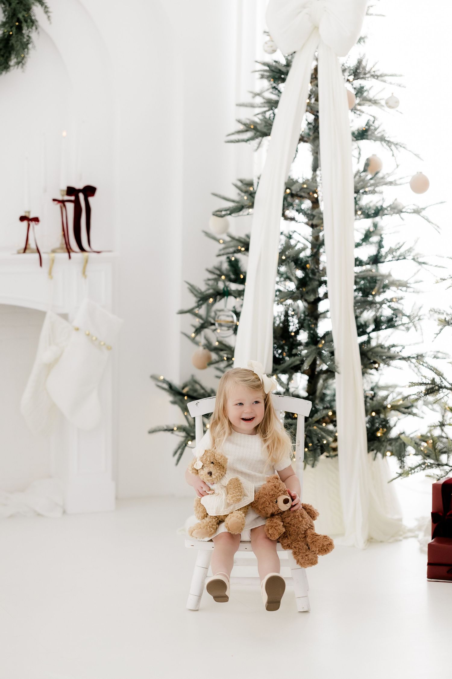 Happy toddler girl in white dress holding teddy bears on chair near decorated Christmas tree in bright white room.