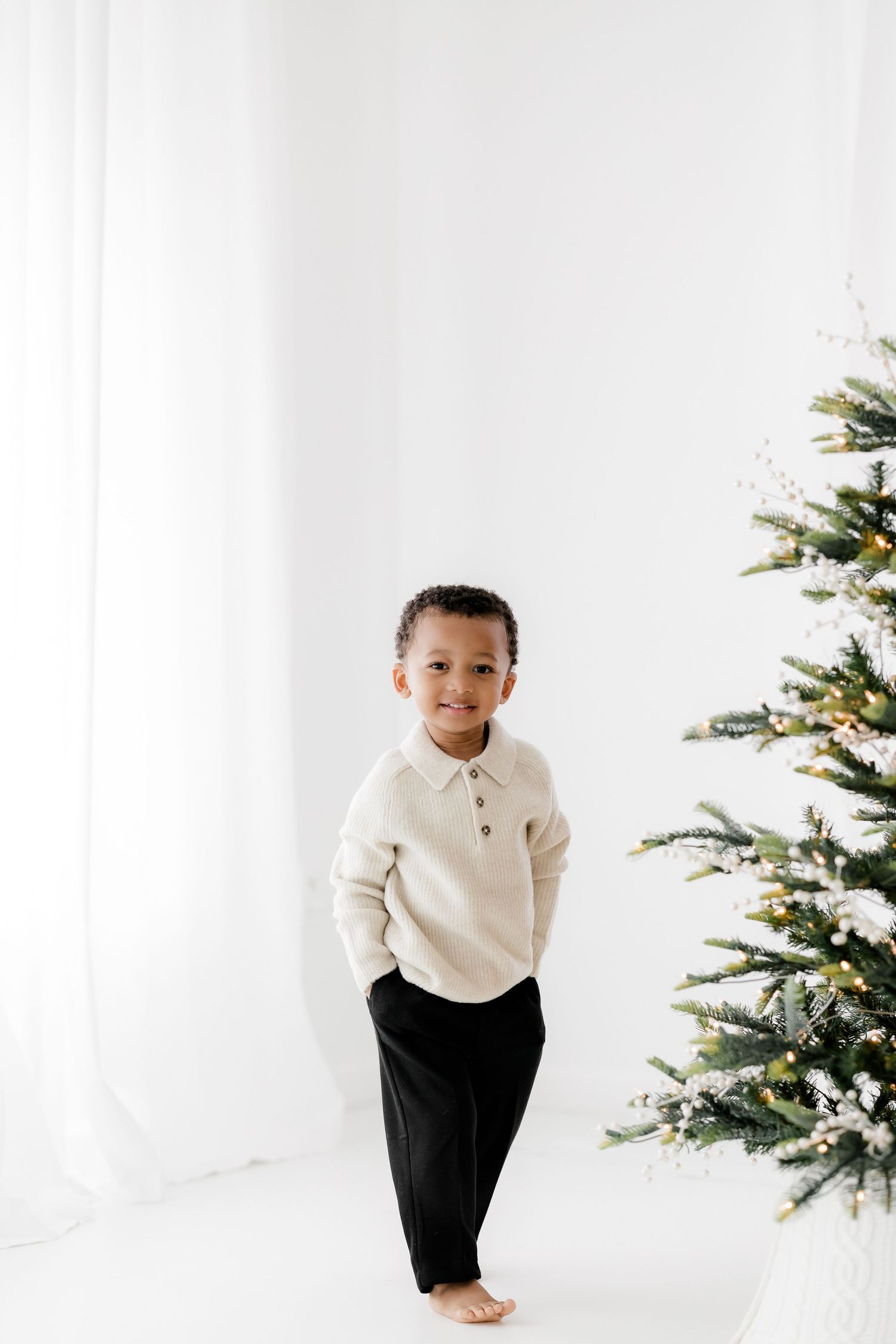 A smiling young boy in a cream sweater and black pants stands barefoot beside a decorated Christmas tree in a bright white room.