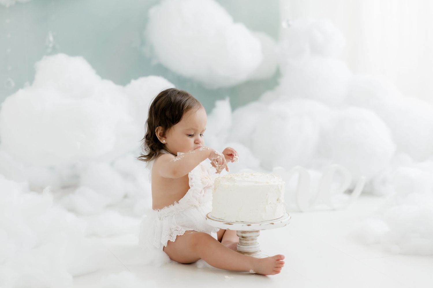 Baby girl in white romper explores a white cake during a dreamy cloud-themed first birthday cake smash photo session.