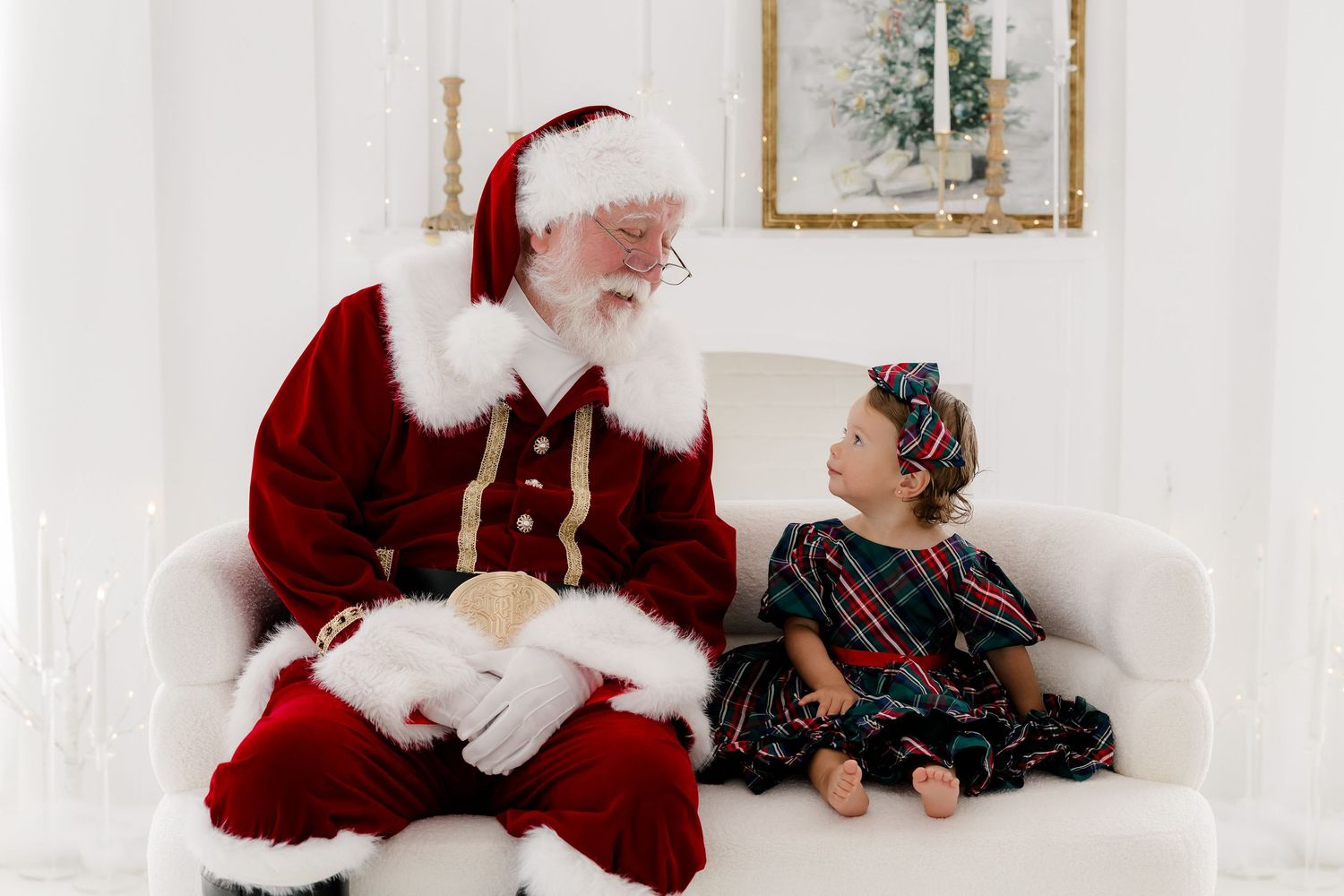 Santa Claus sits on a white sofa with a young girl in a plaid dress during a festive Christmas photo session.