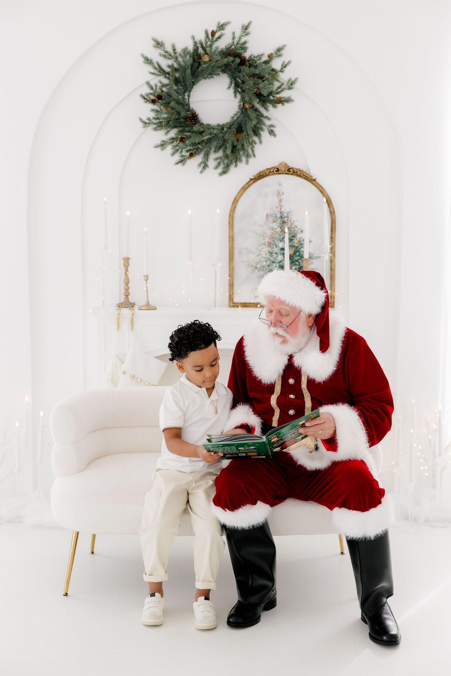Santa Claus reads a Christmas book with a young boy on his lap in a white festive room with wreath decor.