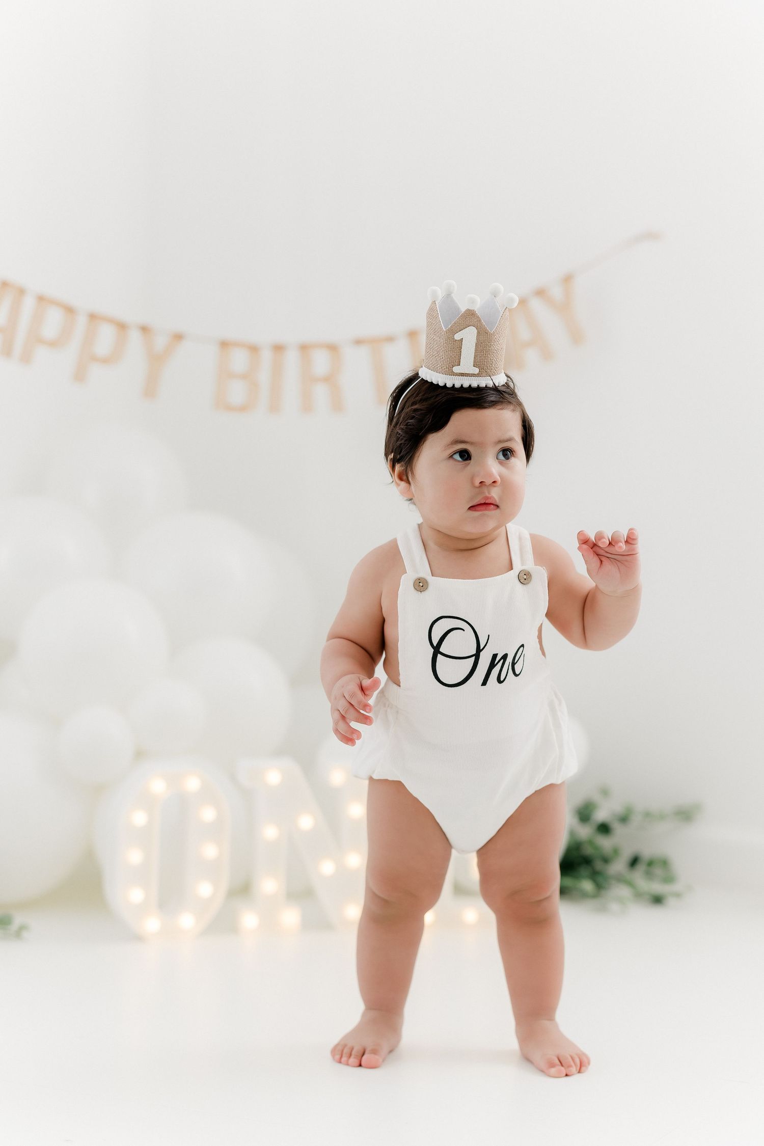 Baby wearing white 'One' onesie and silver crown stands against white backdrop.