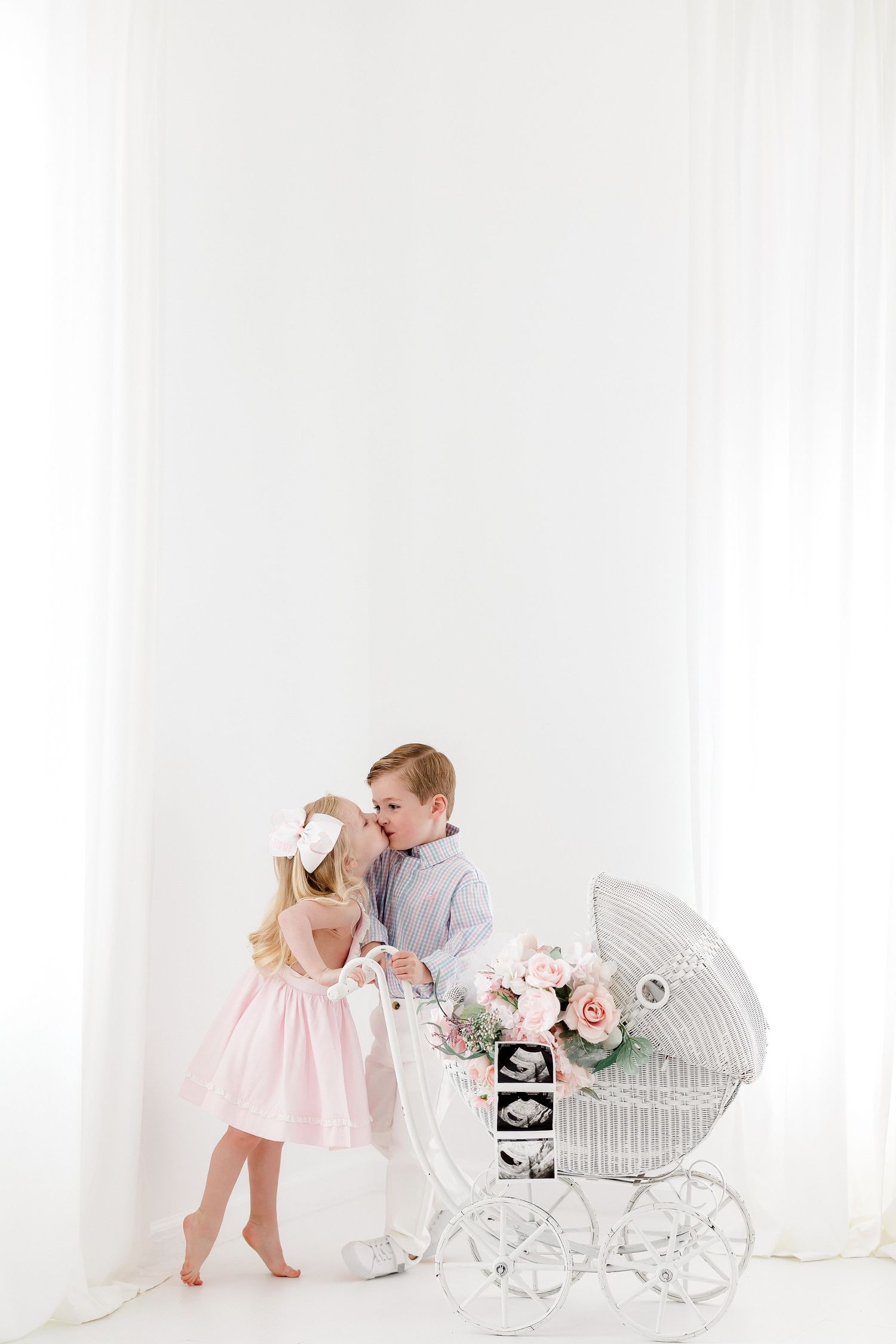 A young boy and girl in pink dress stand beside a vintage white wicker carriage decorated with roses and ultrasound photos.
