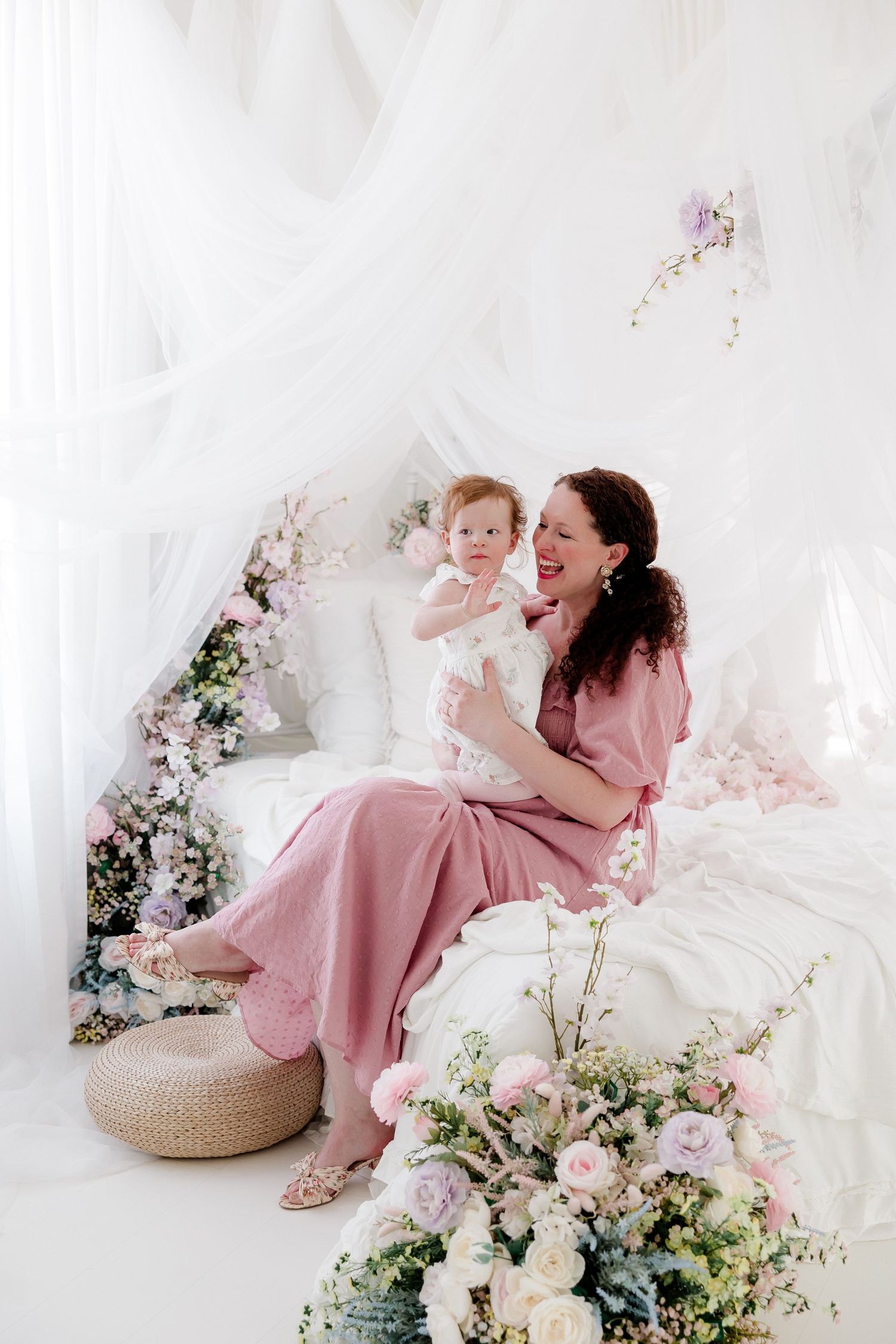 A mother and child share tender moments in a dreamy white studio setting with soft pink dresses and delicate floral arrangements.