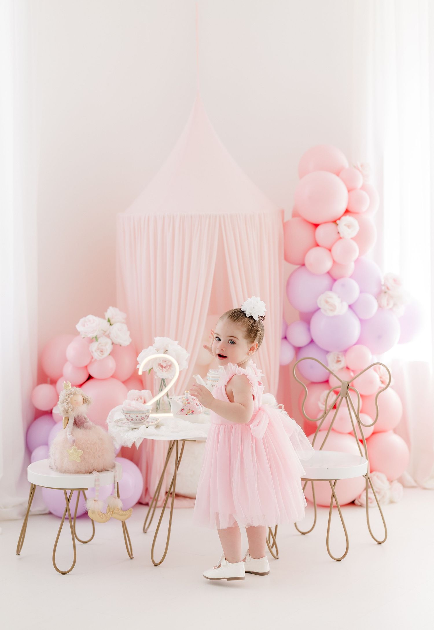 A pink and pastel themed first birthday photo session setup with balloons, floral decorations, and white chairs.