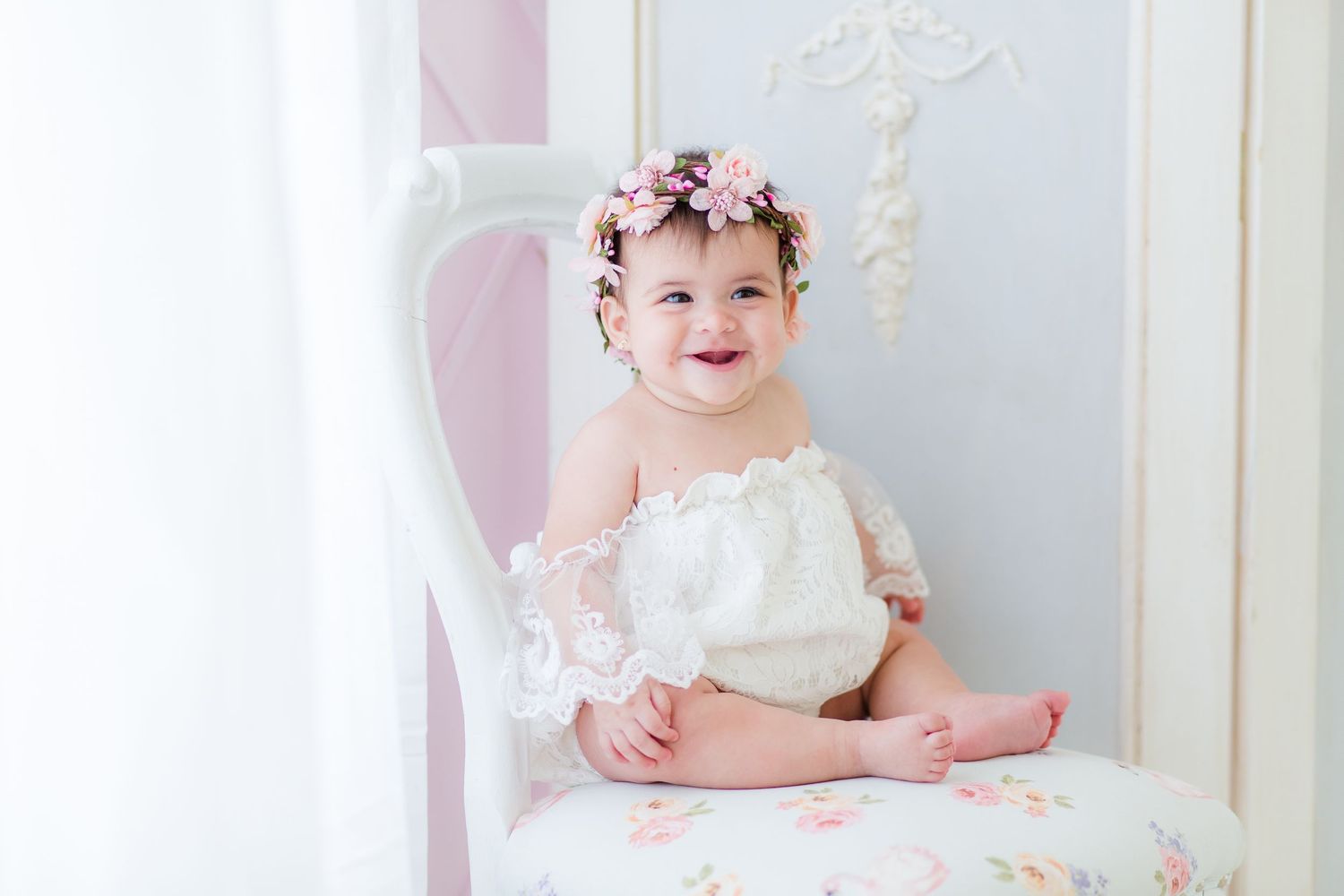 A baby in a white dress sits on a cream-colored couch with pink and white decorative elements in the background.