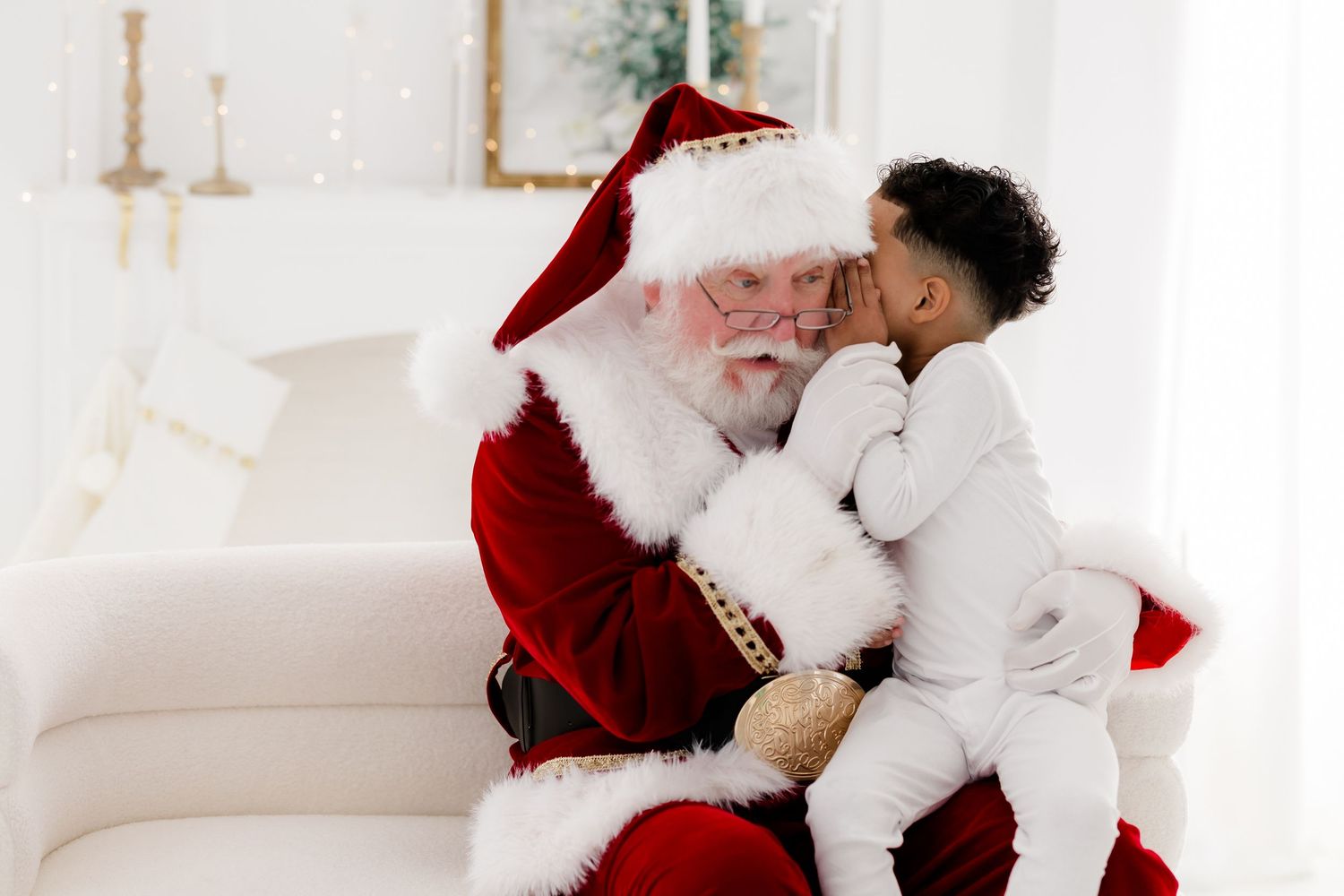 A child whispers to Santa Claus while sitting on his lap in a festive white holiday setting.