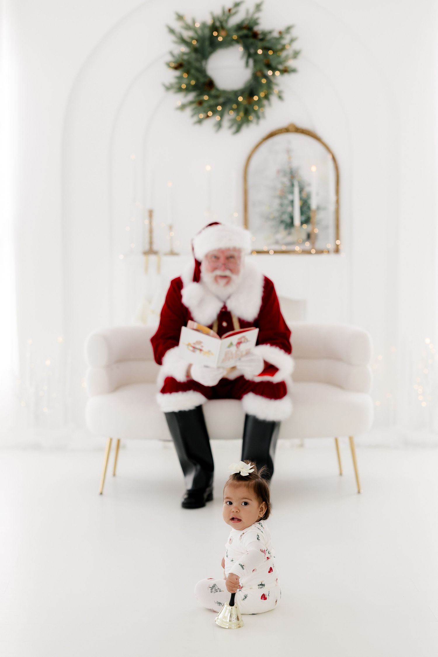 Santa Claus reading a book on a white sofa while a toddler stands in front in a festive holiday studio setting.