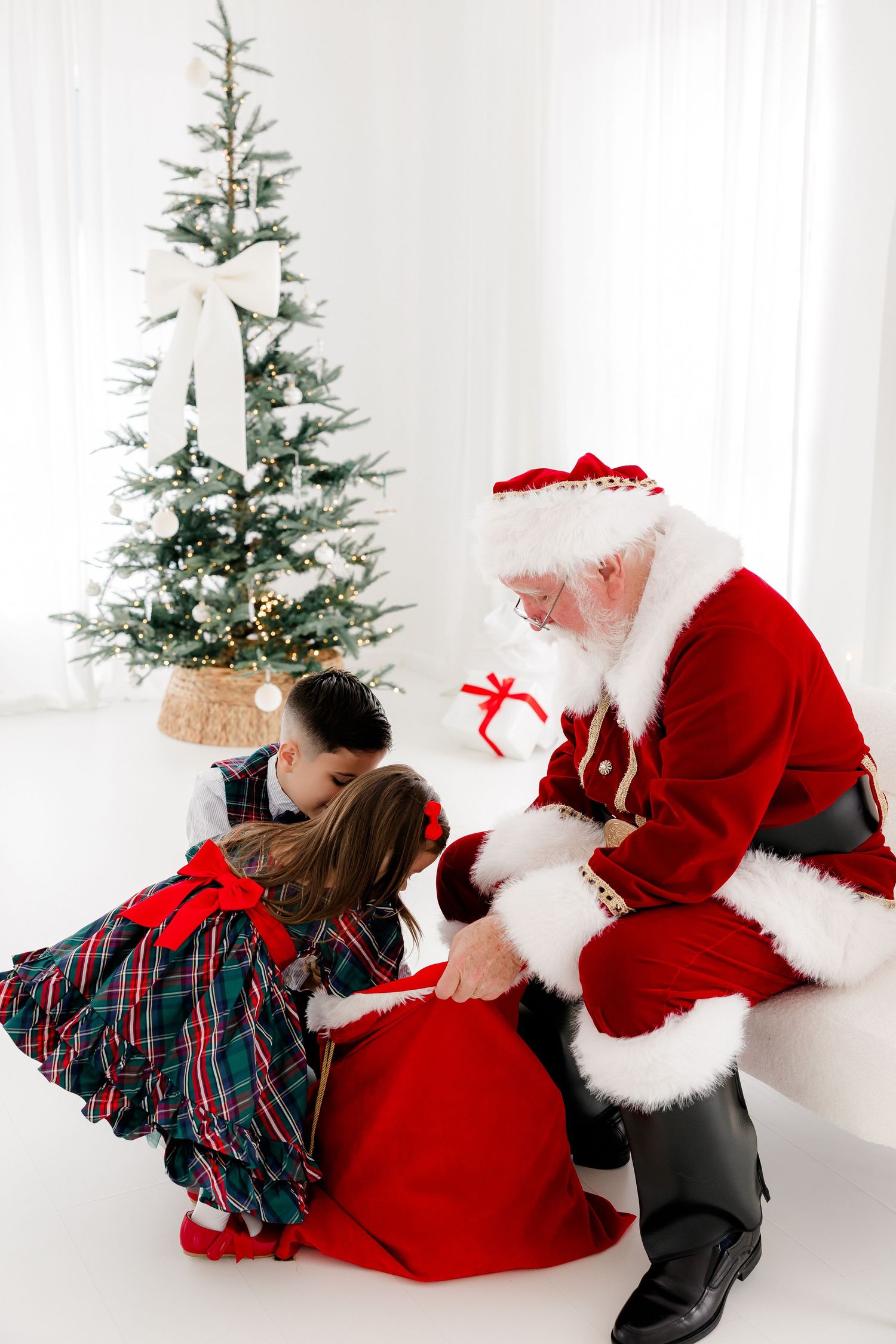 Santa Claus giving gifts to two children by a decorated Christmas tree in a bright white room.