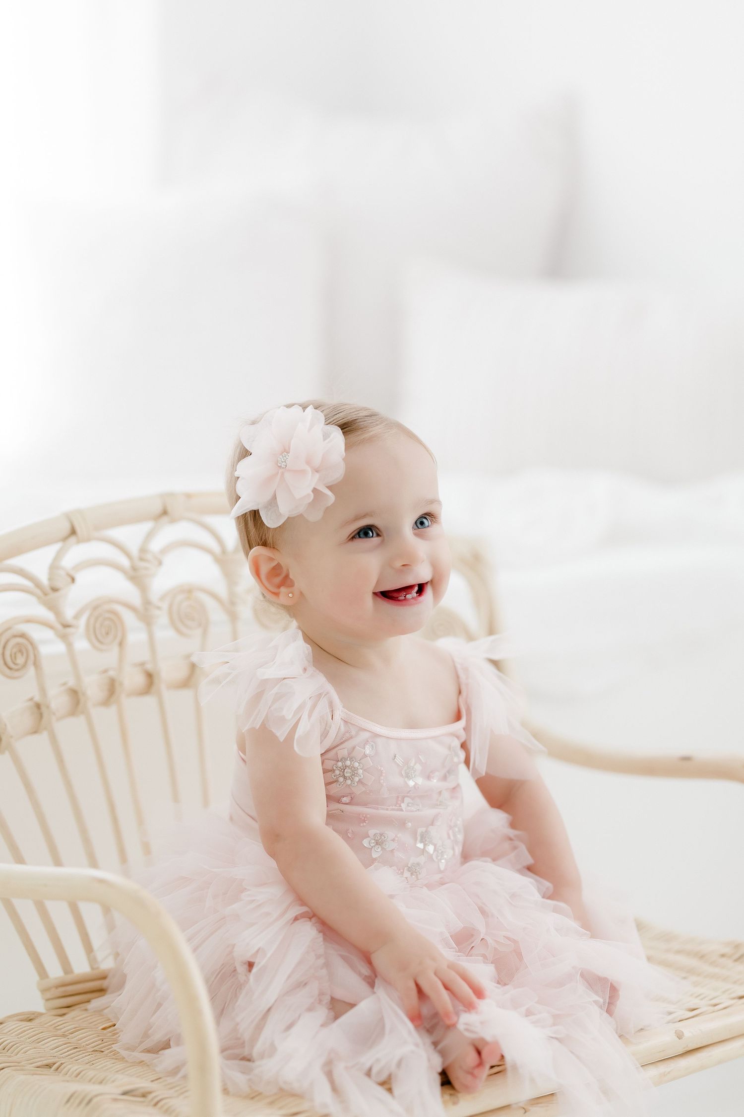 Baby wearing pink tulle dress and floral headband sits smiling in white wicker chair against light background.