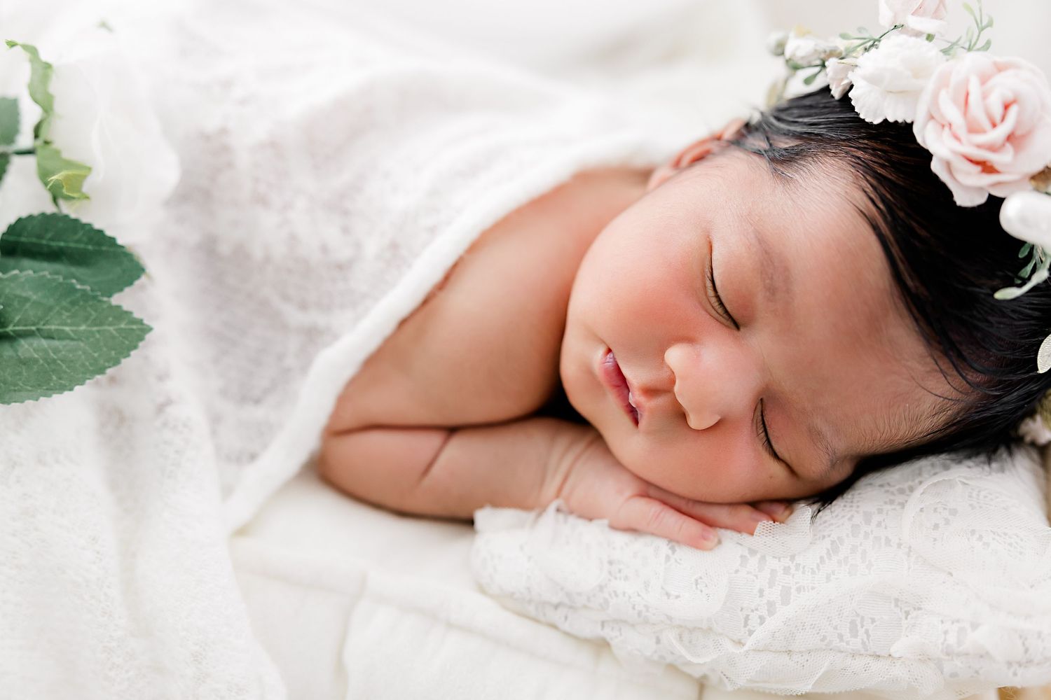 Newborn baby sleeping peacefully on white lace blanket with delicate floral headpiece and soft lighting.
