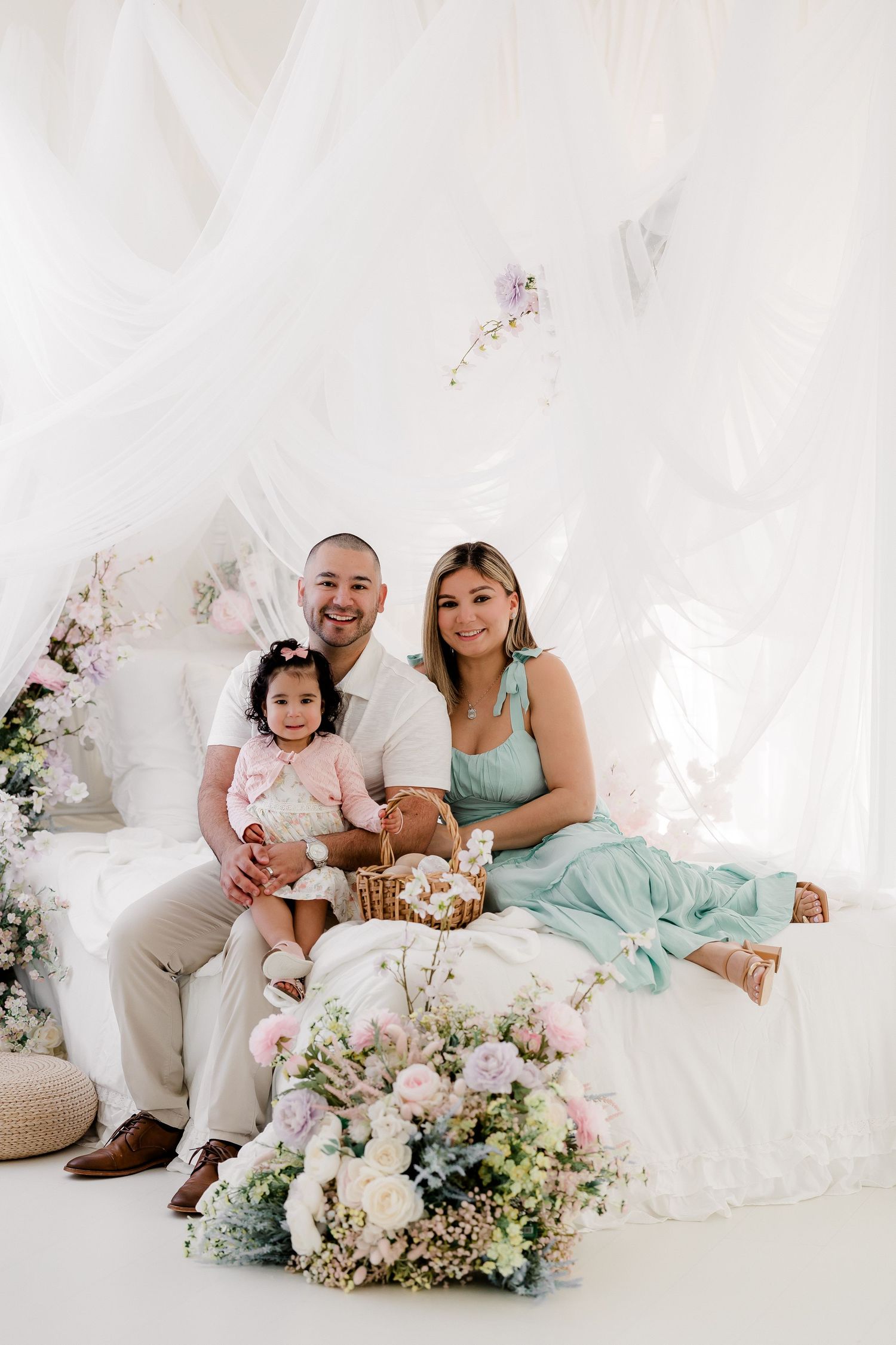 Family poses together on white couch with floral decorations and soft drapery in dreamy studio setting.