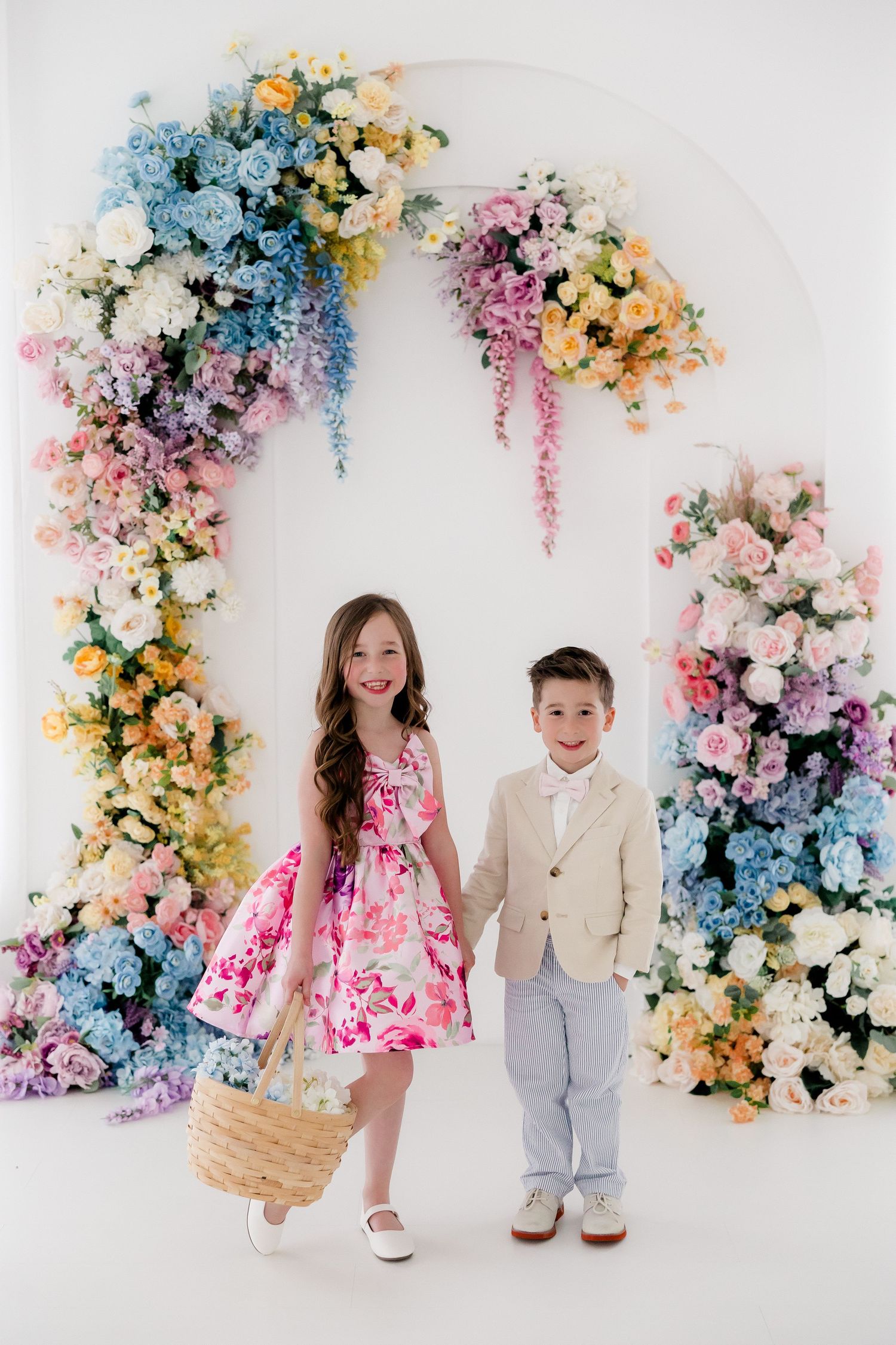 Children pose together against a stunning floral arch backdrop with pink, blue, and yellow blooms in a bright studio setting.