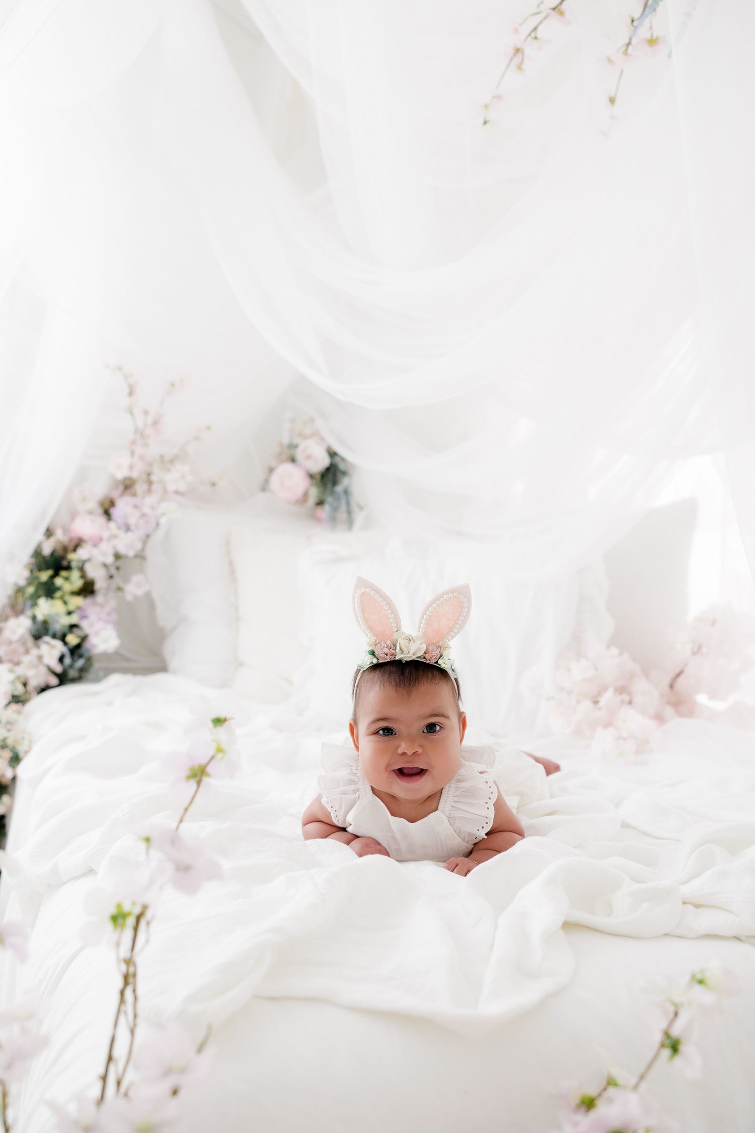 A baby in white clothing lies on soft white bedding surrounded by delicate flowers and ethereal decor.