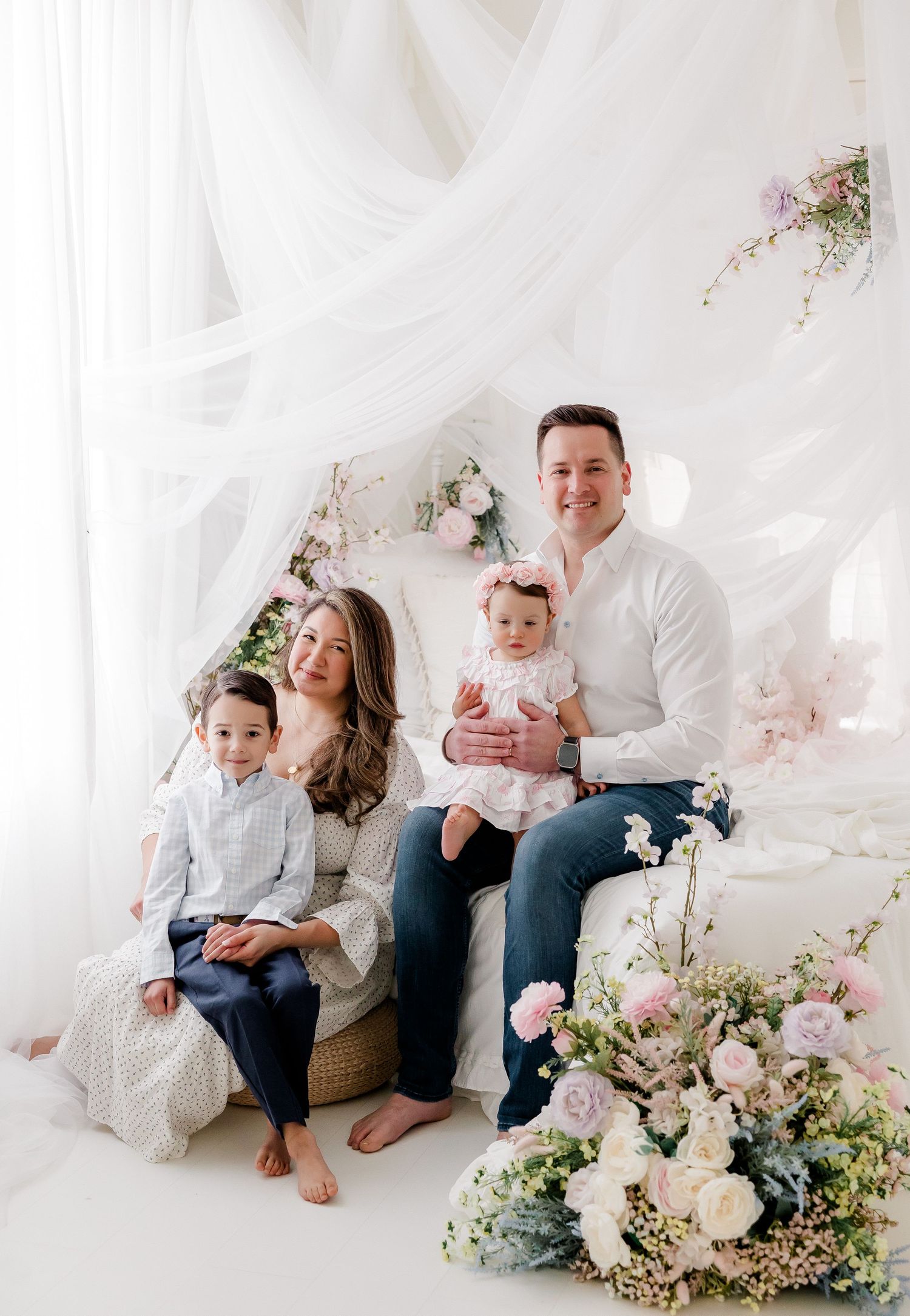 Family portrait in white studio setting with flowing fabric backdrop and floral arrangements creating an ethereal atmosphere.