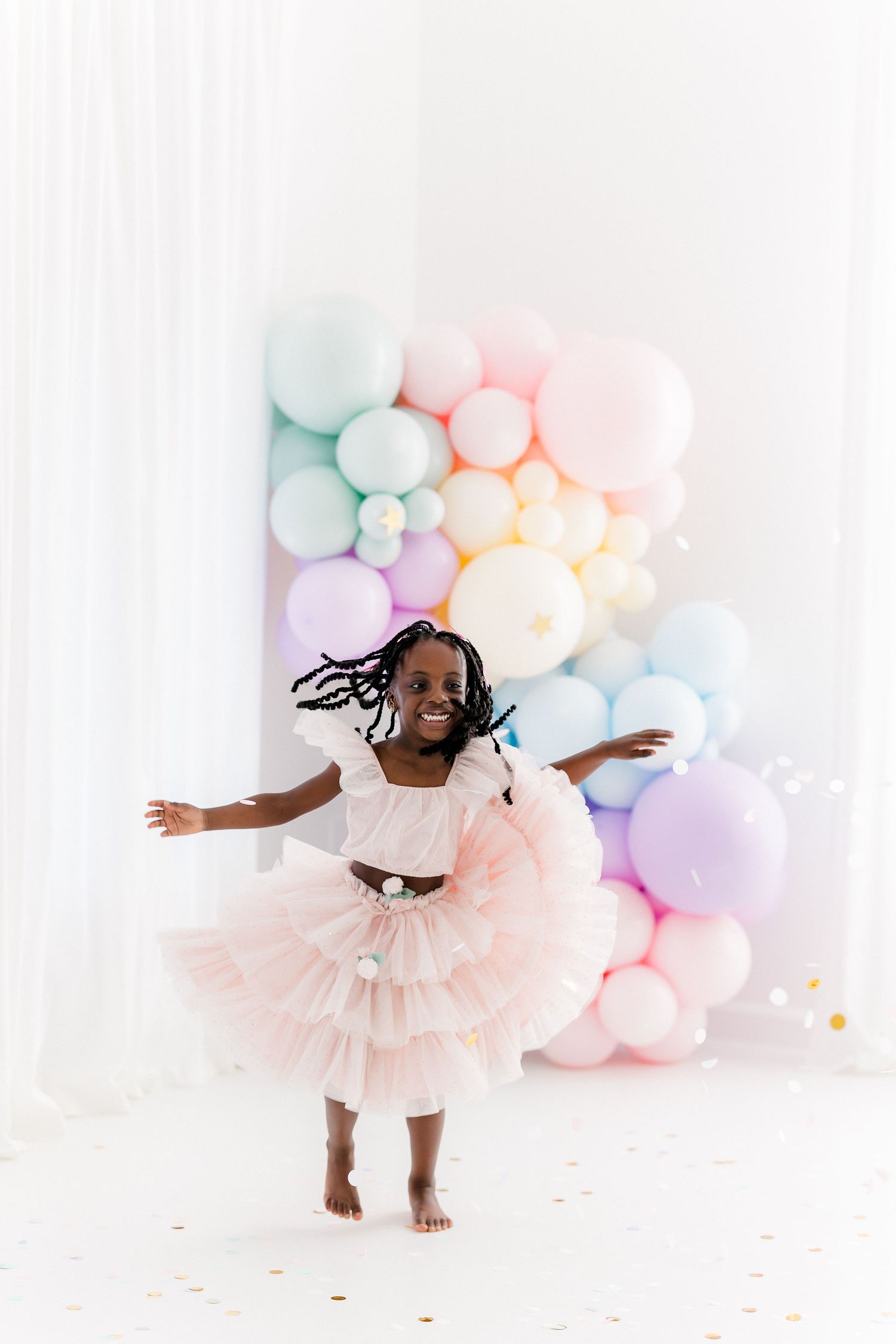A toddler in a white tulle dress twirls and dances in front of a pastel balloon arch backdrop.