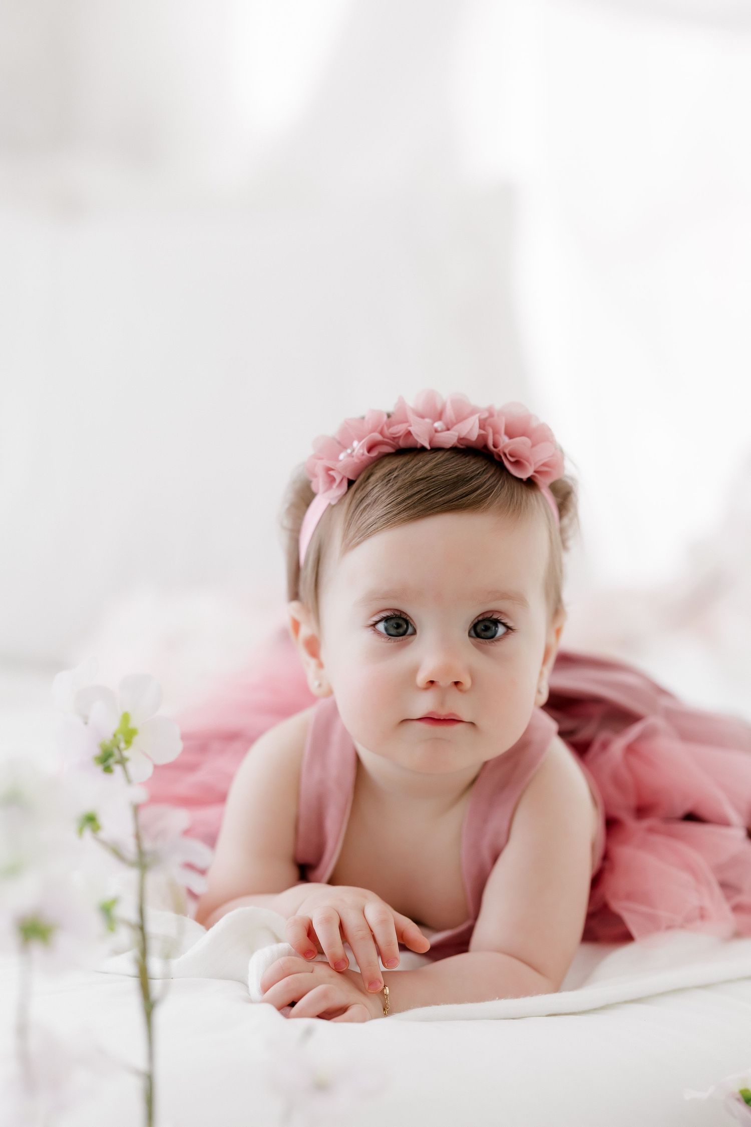 A baby in pink dress laying on white bedding with delicate floral elements.